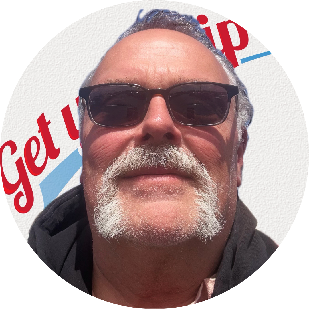 Close-up of a man with gray hair, a mustache, and beard wearing sunglasses, standing in front of a white background with the partial words 'Get u' visible.