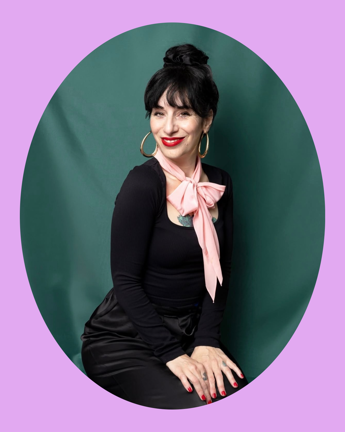 A woman with black hair styled in a bun, wearing large gold hoop earrings, a black long-sleeve top, a pink neck scarf tied in a bow, and black pants, sitting against a green background with a purple frame.