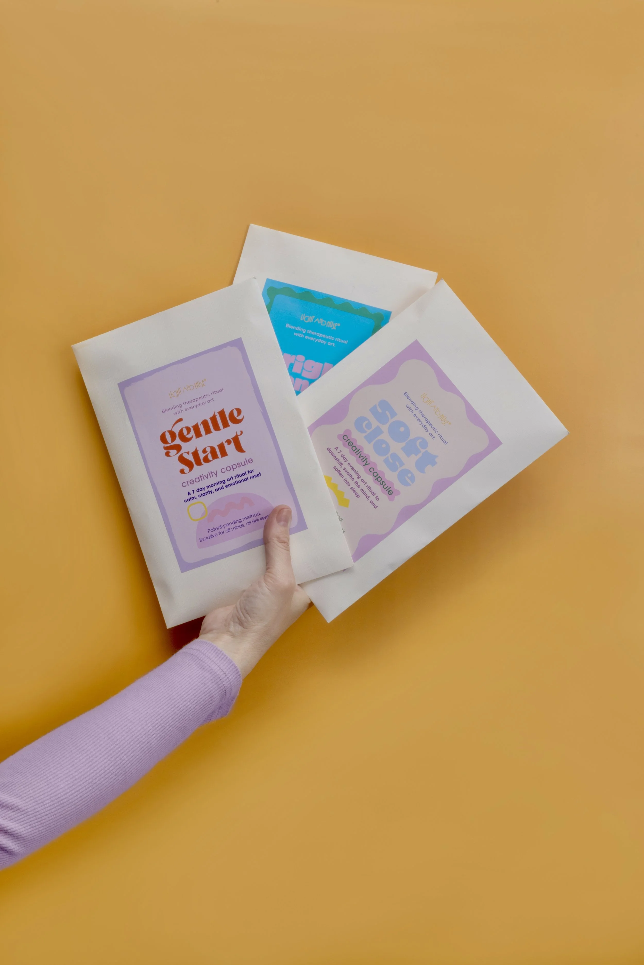 A hand holding three colorful packages on a yellow background. The packages are labeled 'Gentle Start,' 'Creative Cap,