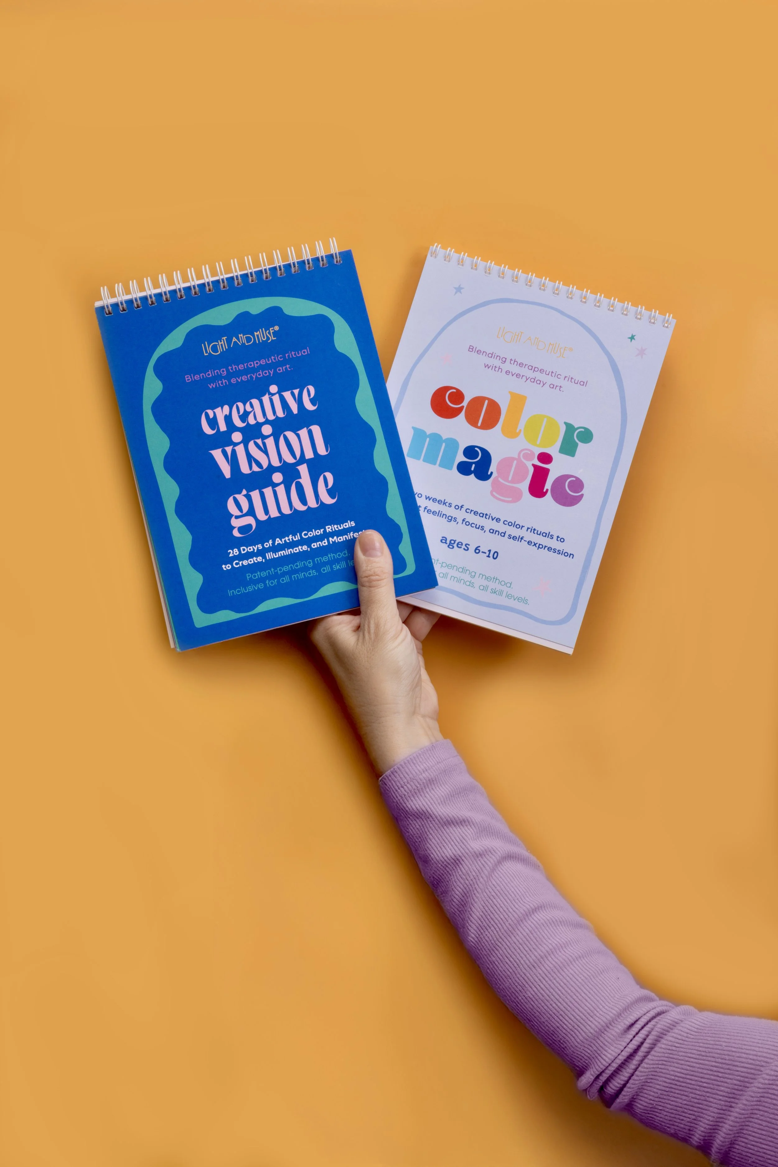 Two spiral-bound booklets about art therapy and color magic are held against an orange background. The booklet on the left is blue and titled 'Creative Vision Guide,' with purple text and a colorful wavy border. The booklet on the right is white and titled 'Color Magic,' with rainbow-colored text and small pastel stars.