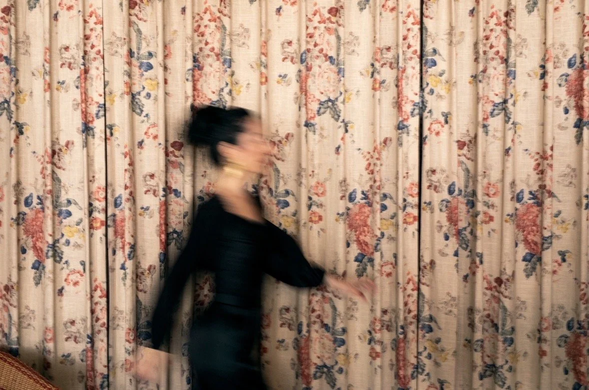 A blurry woman with dark hair in a bun wearing a black dress walking past floral-patterned curtains.