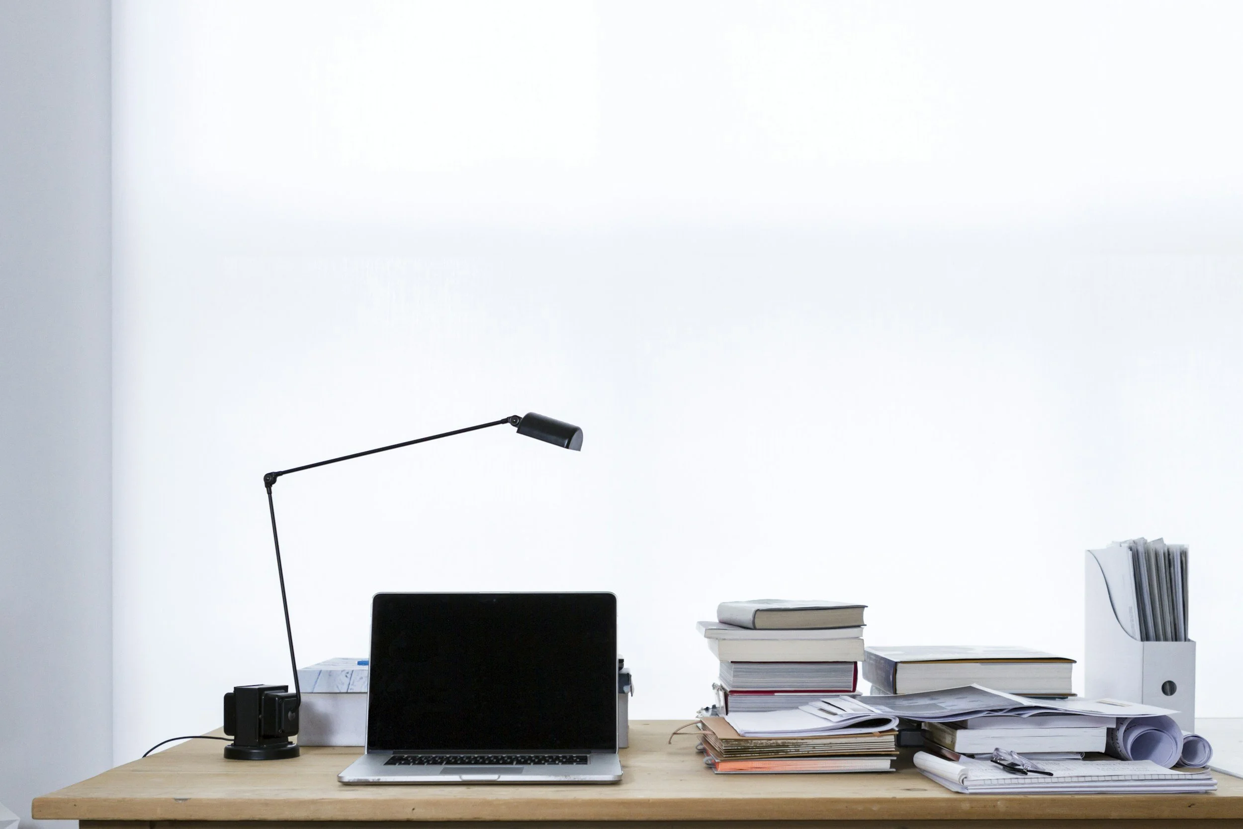A cluttered wooden desk with a laptop, stacked books, scattered papers, a desk lamp, a black speaker, and rolled-up papers against a plain white wall.