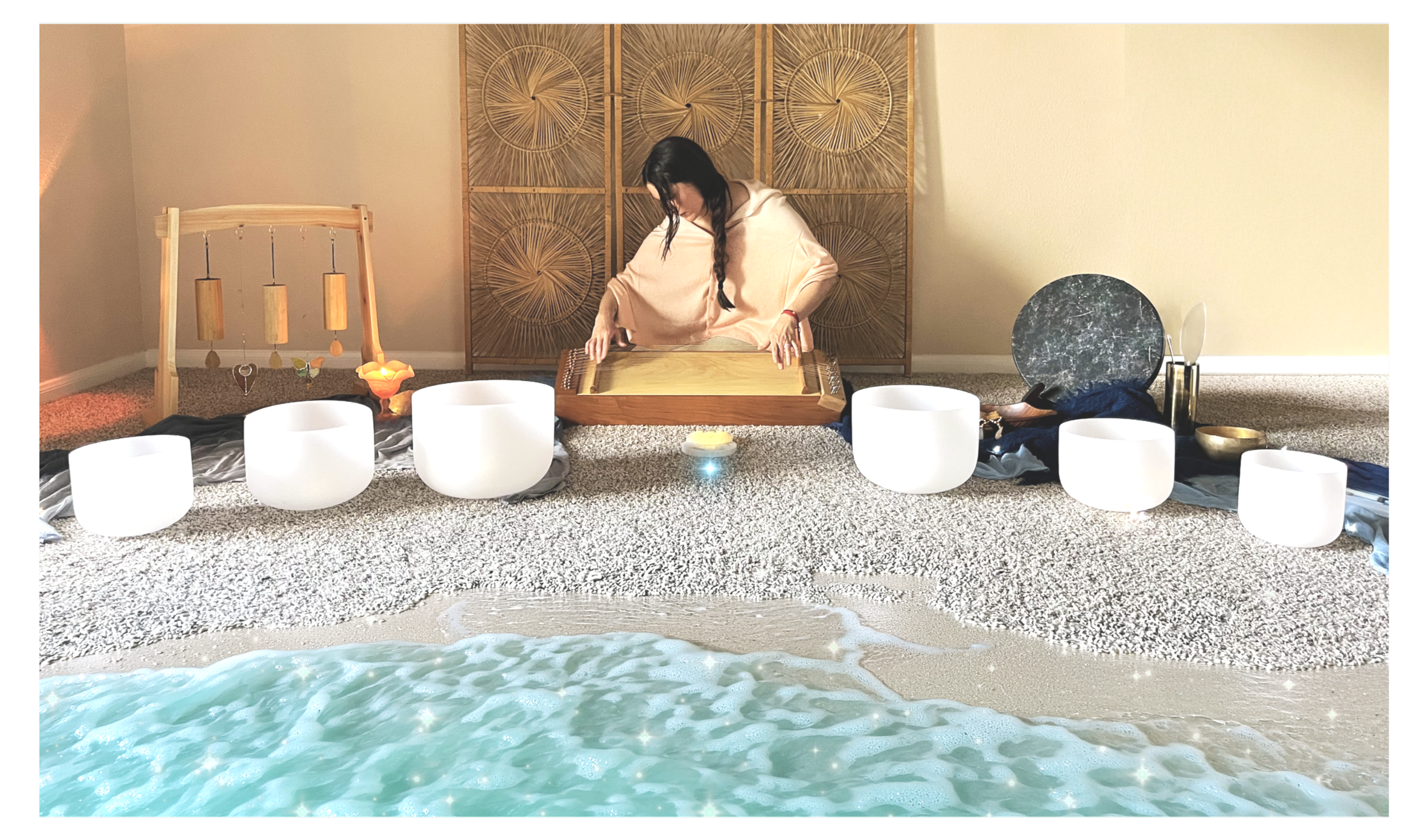 A woman with dark hair in a braid wearing a pink gradient cape is sitting cross-legged on a textured carpet, playing a stringed instrument in a room with white singing bowls and soft lighting, with a safe gentle wave in the foreground.