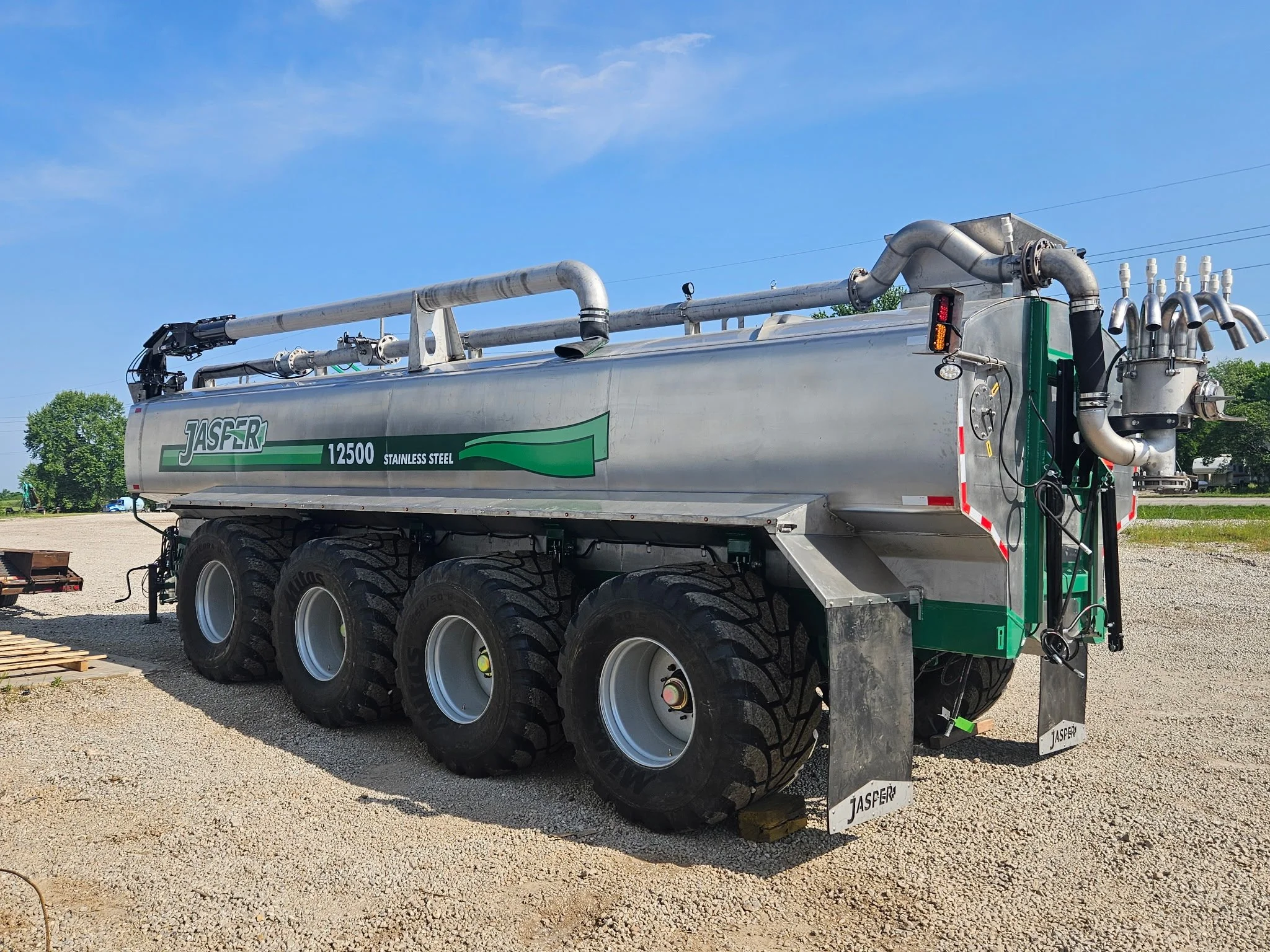 A large stainless steel tanker truck with black tires and green accents, labeled 'Jasper 12500,' is parked outdoors on a gravel lot under a blue sky with some clouds. The truck has multiple pipes and equipment on top, and the back of the truck featur