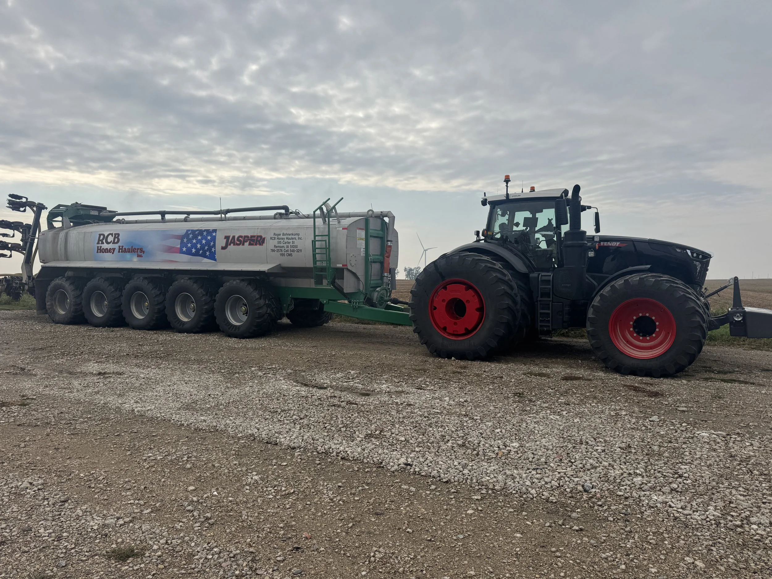 A large tractor connected to a tanker trailer on a gravel field under a cloudy sky. The tanker displays branding for RCB Honey Haulers and Jasper, with an American flag design.