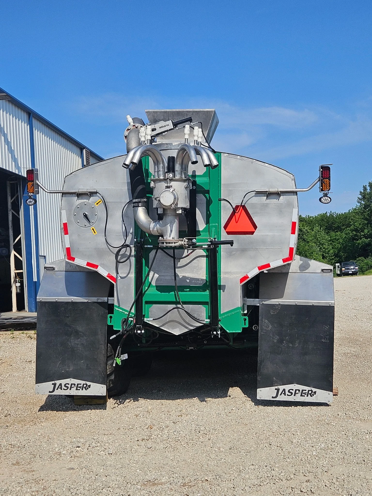 Recycling or waste management tanker truck with 'Jasper' brand name, parked outdoors on a gravel surface next to a building with a blue roof, under a clear blue sky.