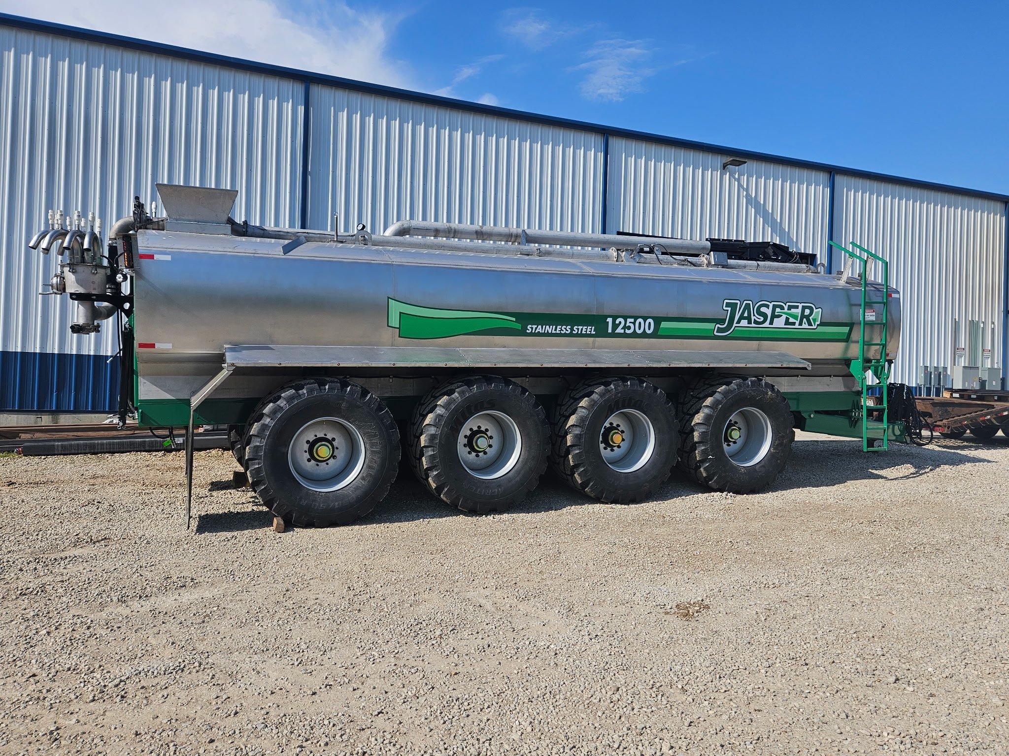 A large stainless steel tanker truck with green and white branding sitting on gravel ground in front of a blue and white industrial building under a partly cloudy sky.