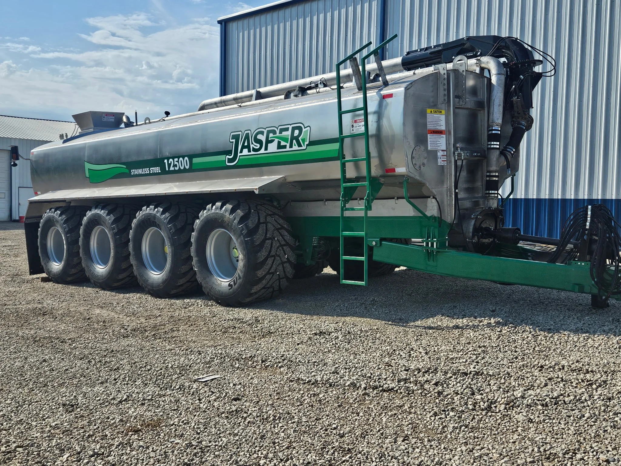 Large stainless steel tanker truck with green and white branding on a gravel lot next to a blue and silver industrial building, with a partly cloudy sky above.