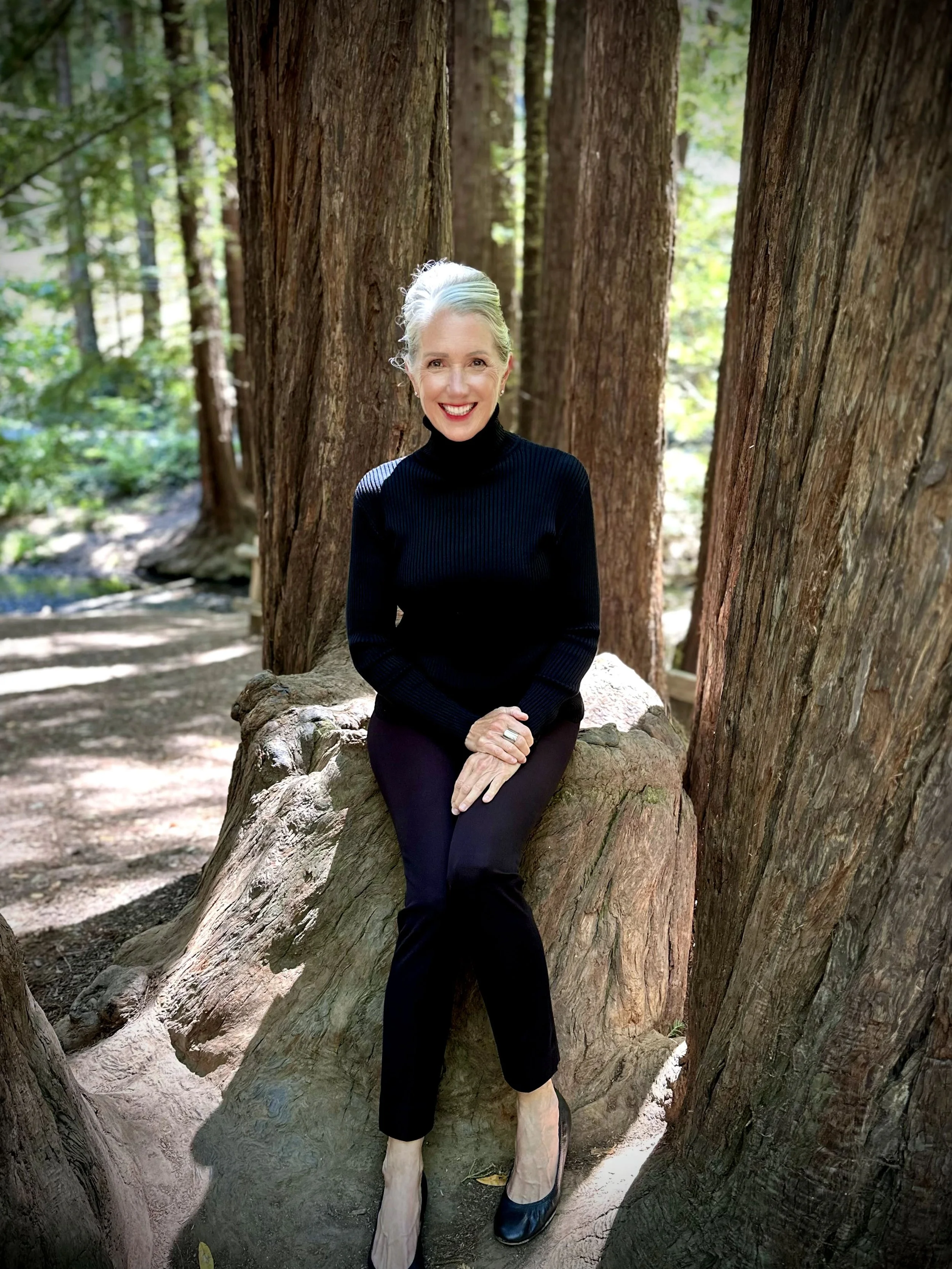 A woman with gray hair, wearing a black turtleneck and black pants, sitting on a large tree root, smiling in a forest with tall trees and greenery in the background.