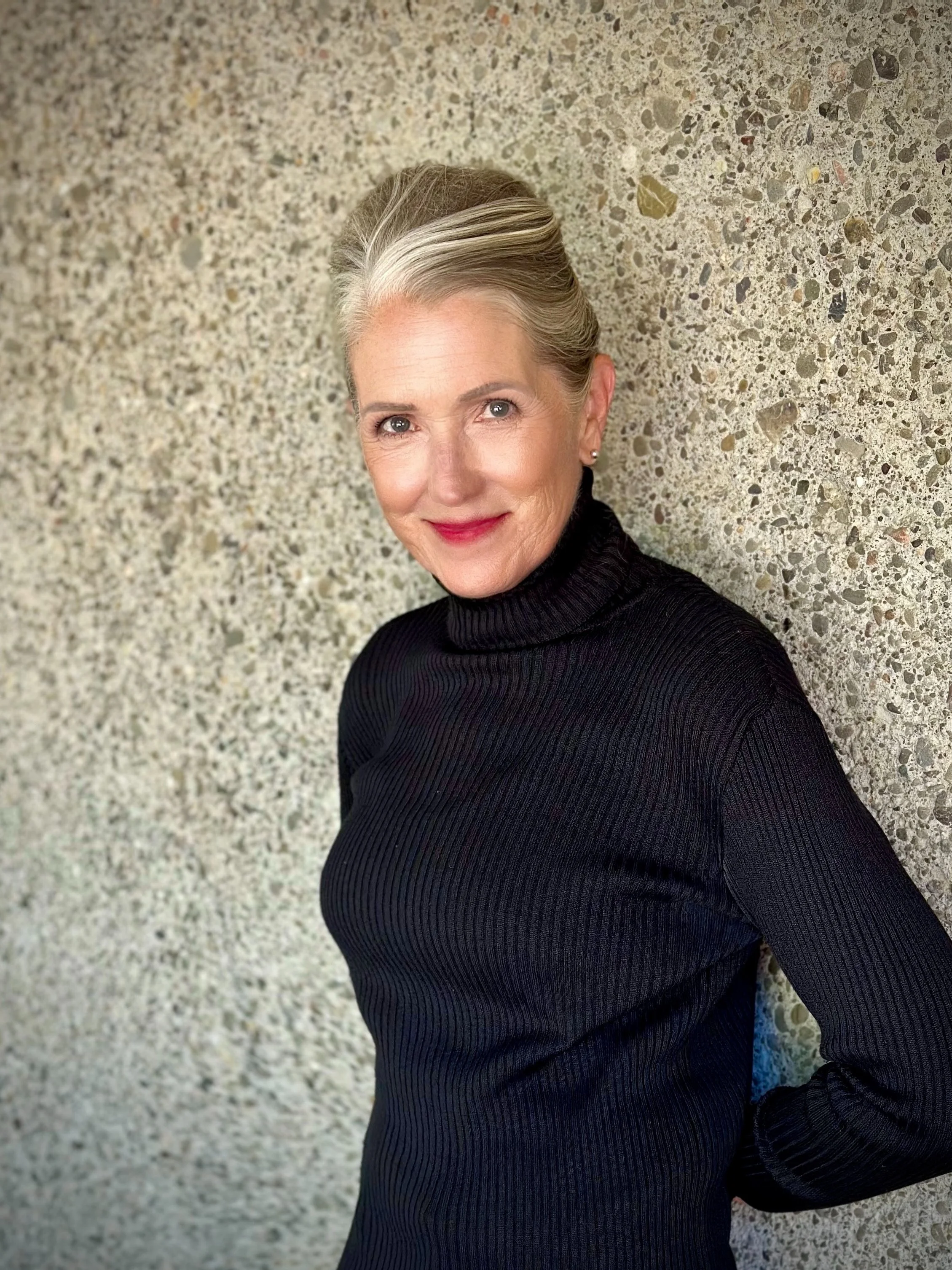 A woman with short gray hair, red lipstick, and earrings, wearing a black turtleneck, leaning against a textured beige wall with her hands behind her back.
