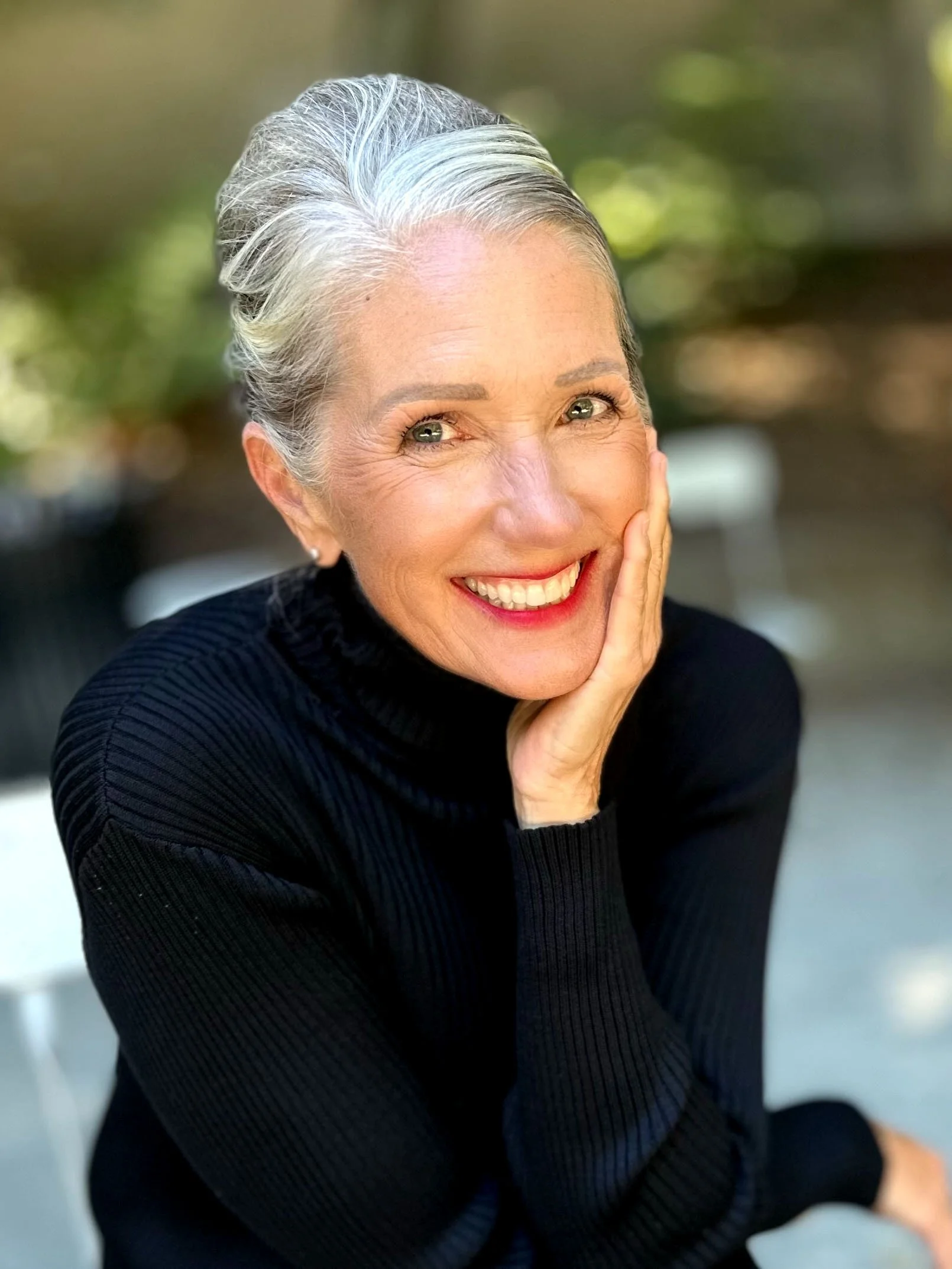 An woman with wearing a black long-sleeve top, smiling with one hand touching her cheek, outdoors with blurred green background.