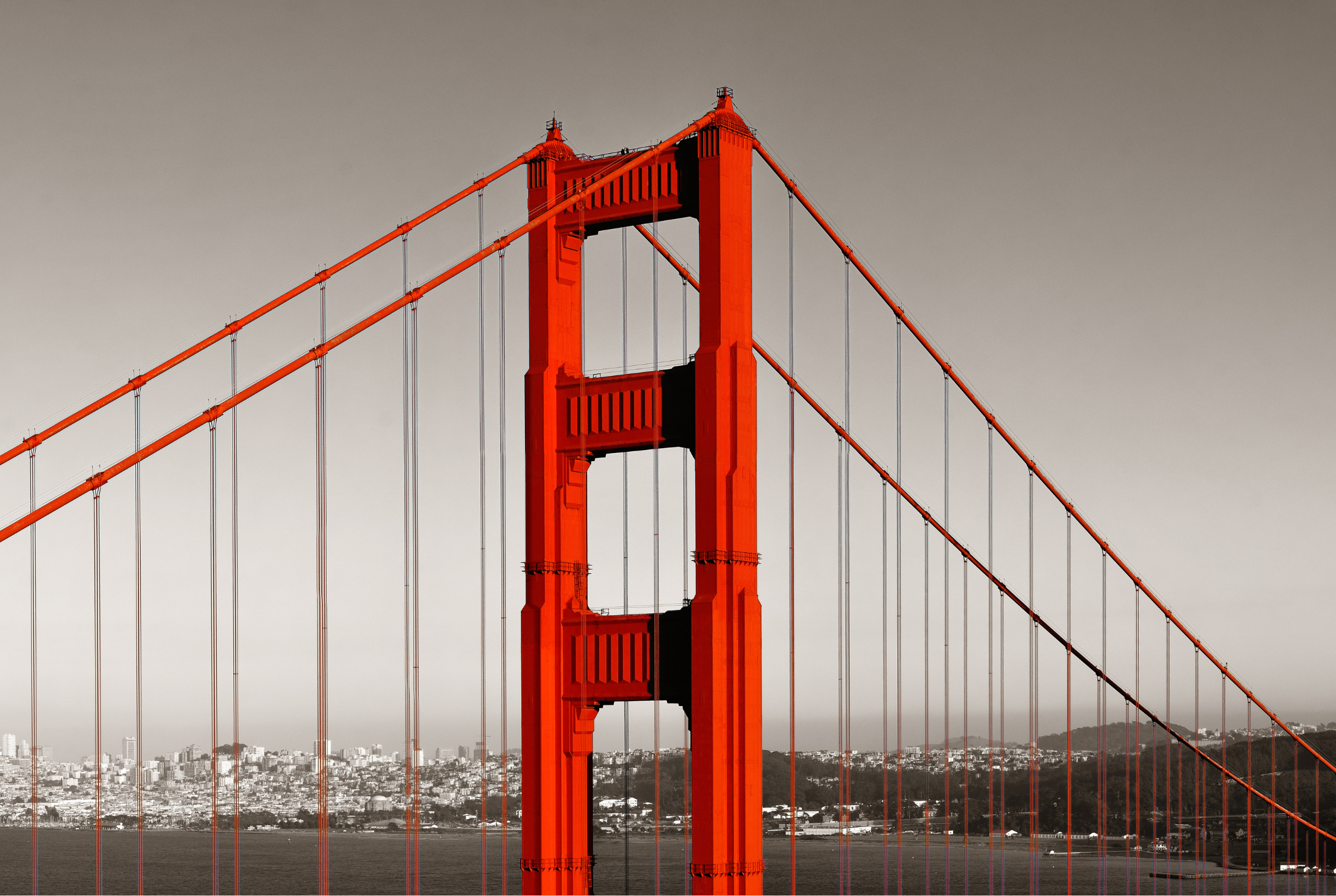 Close-up of the Golden Gate Bridge's orange tower with suspension cables against a gray sky.