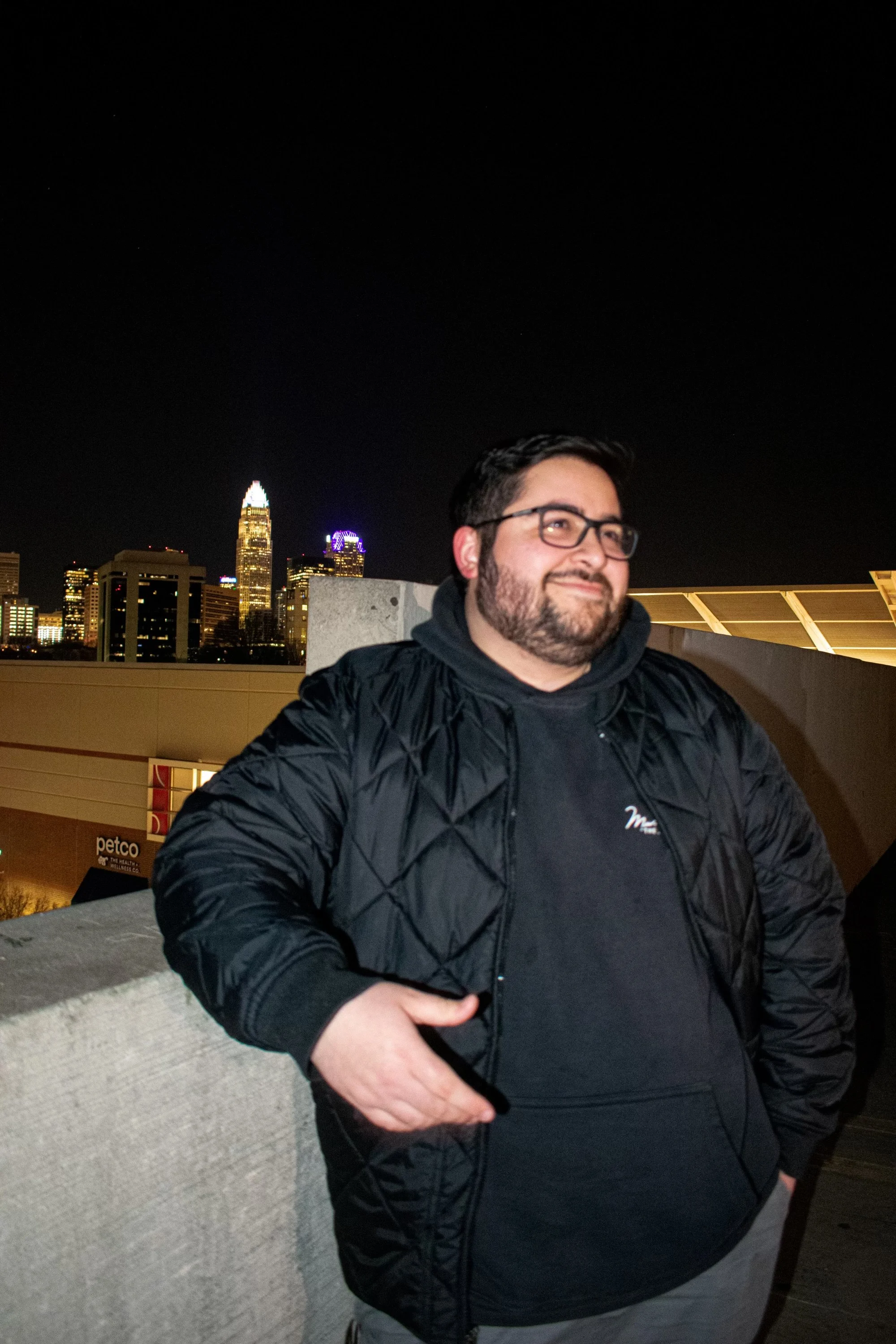 A man with glasses and a beard, smiling, wearing a black jacket and black hoodie, standing on a rooftop with the Charlotte skyline at night in the background.