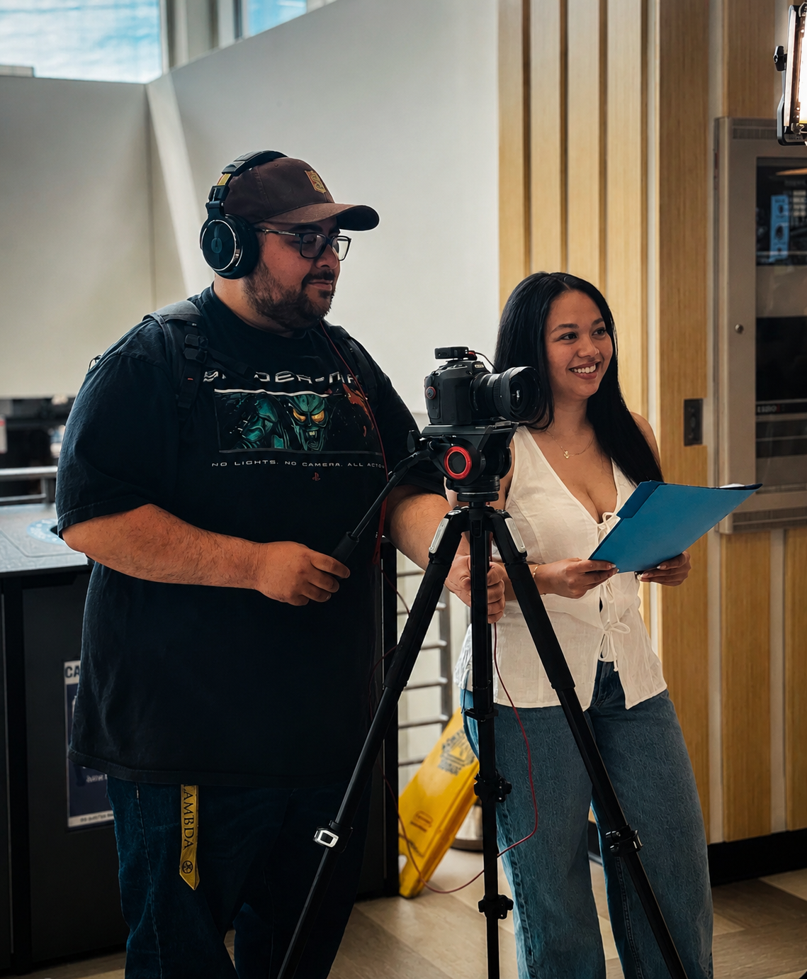 Two people, a man and a woman, setting up video recording with a camera on a tripod in an indoor space.
