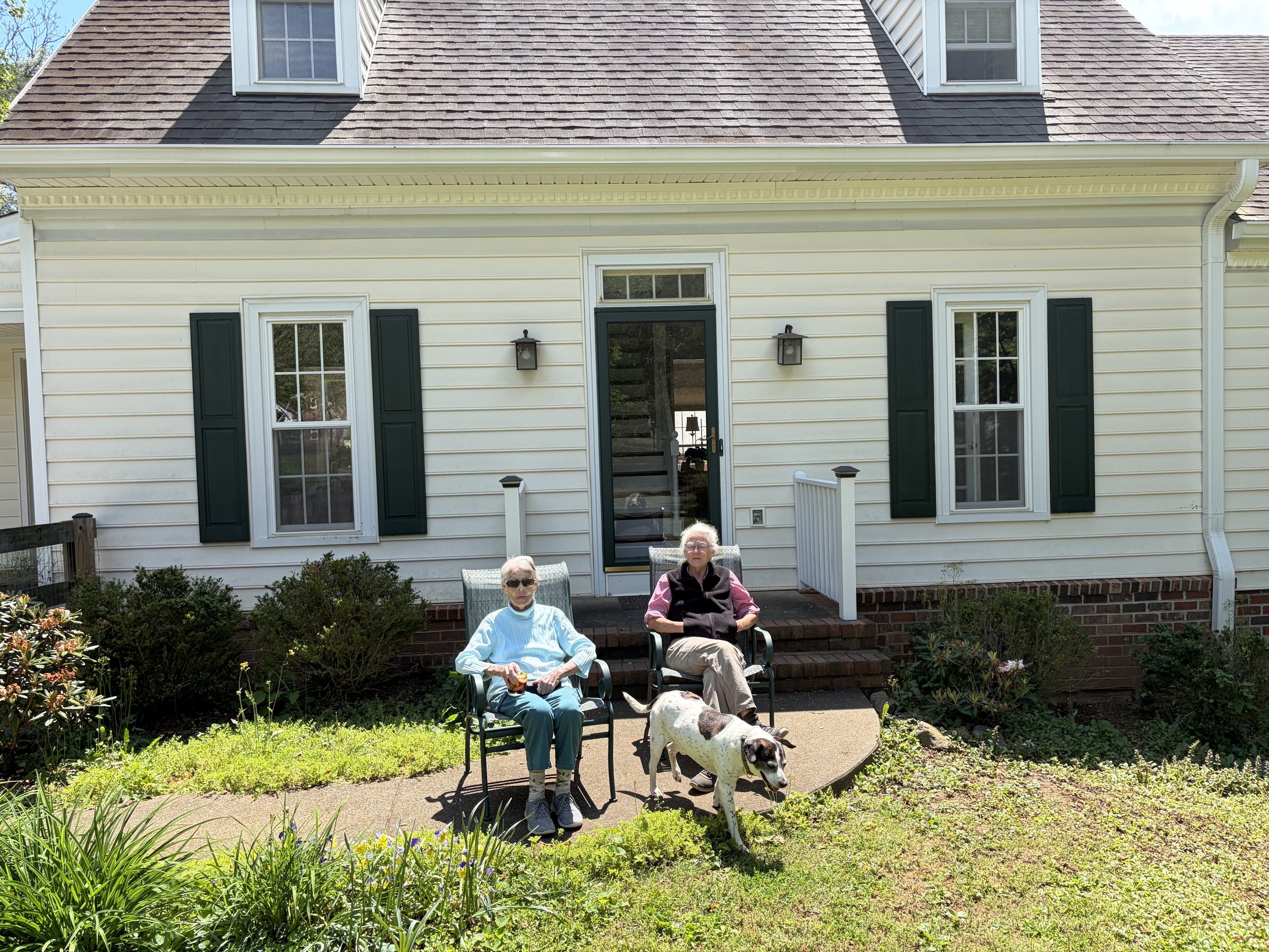 Two elderly women sitting in lawn chairs in a backyard garden with a dog, in front of a yellow house with green shutters and a brick patio