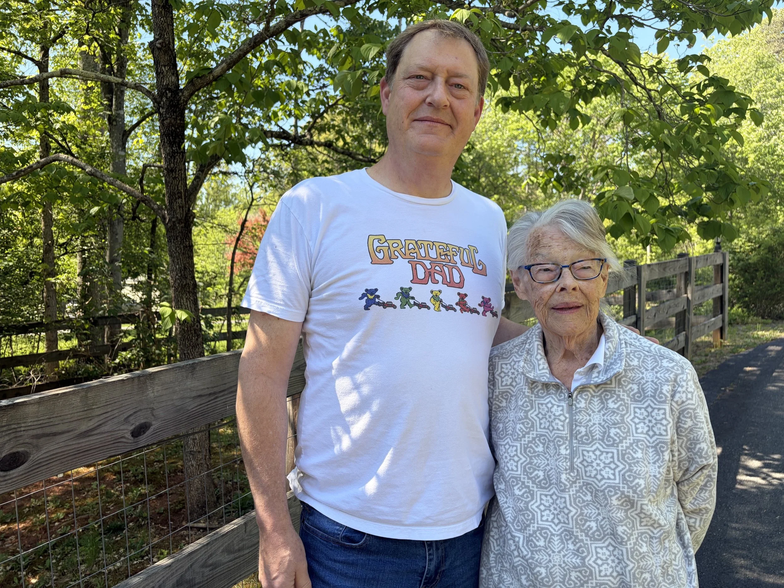 A middle-aged man and an elderly woman standing outdoors on a sunny day, with trees and a wooden fence in the background. The man is wearing a white T-shirt with colorful teddy bear illustrations and text that reads "GREATFUL DAD." The elderly woman has gray hair, glasses, and is wearing a patterned jacket.