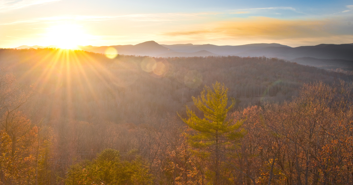 Sun setting over a forested mountain landscape with orange and yellow foliage and a clear sky.