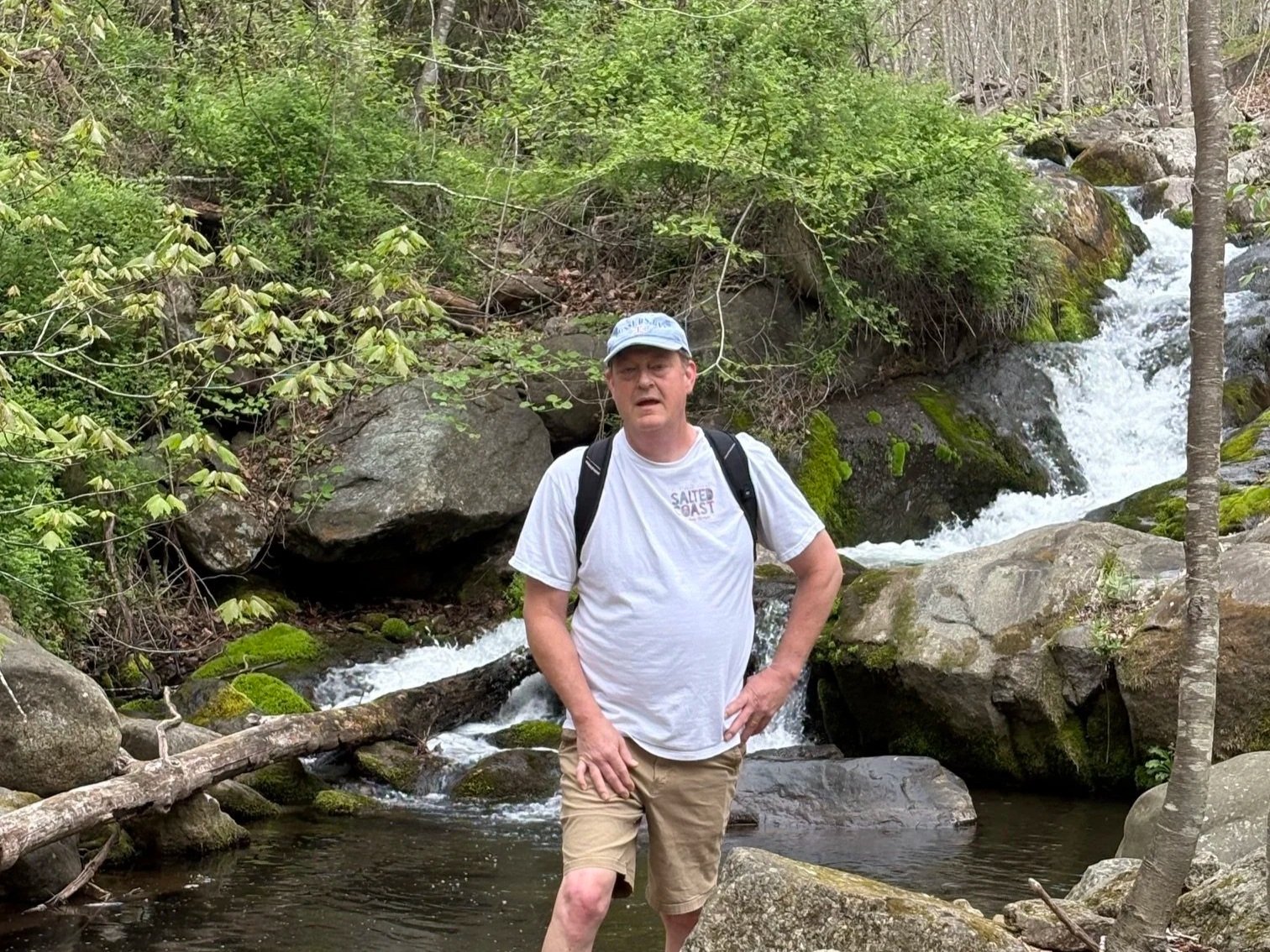 A man in a white t-shirt, tan shorts, and a light blue cap stands in a small creek with a waterfall and rocks in the background, surrounded by green trees and bushes.