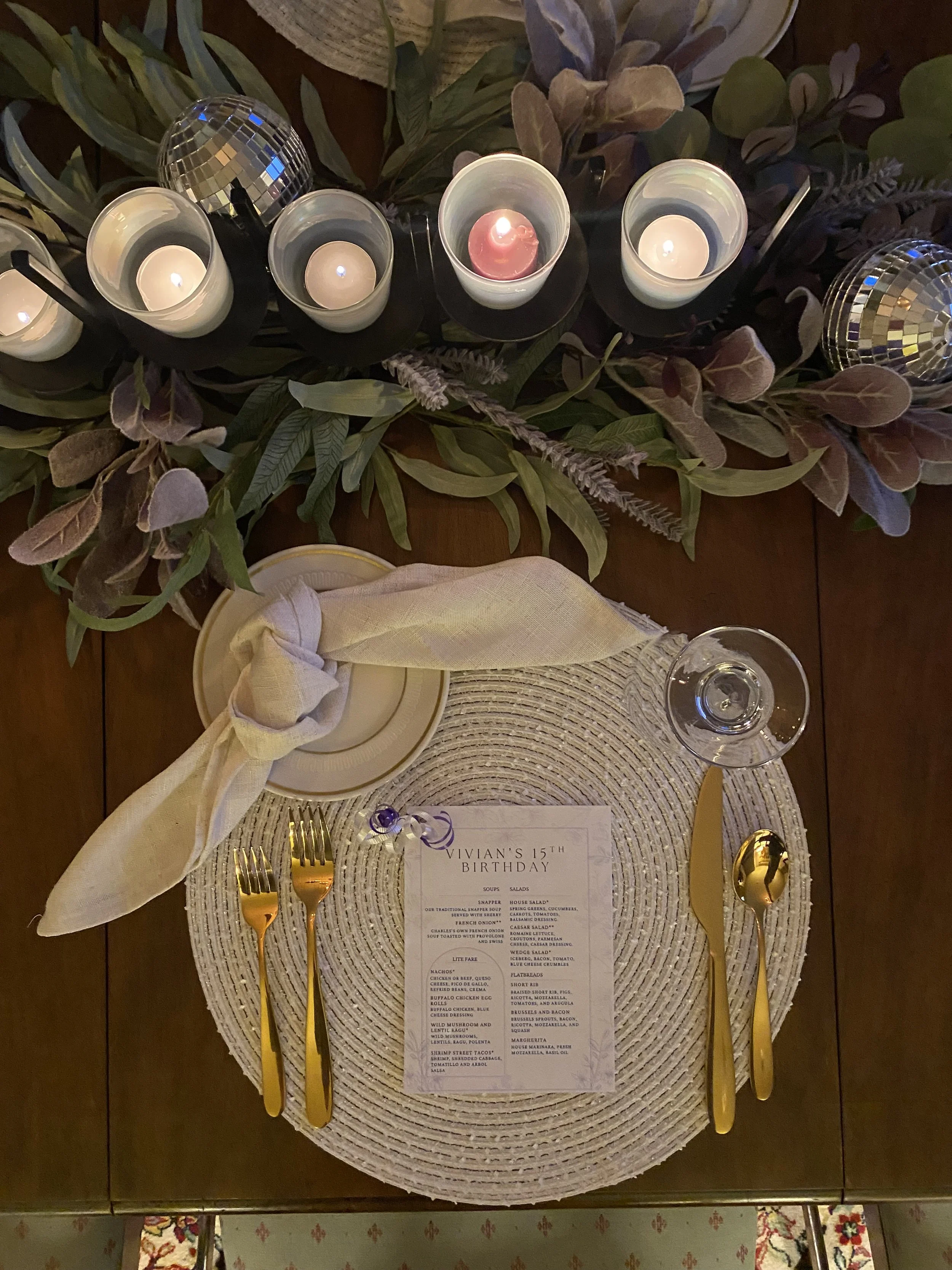 A celebration table setting with a menu card titled 'Vivian's 15th Birthday'. The table includes gold utensils, white plates with yellow napkins, surrounded by greenery and candles, with two disco balls and a lit candle in the center.
