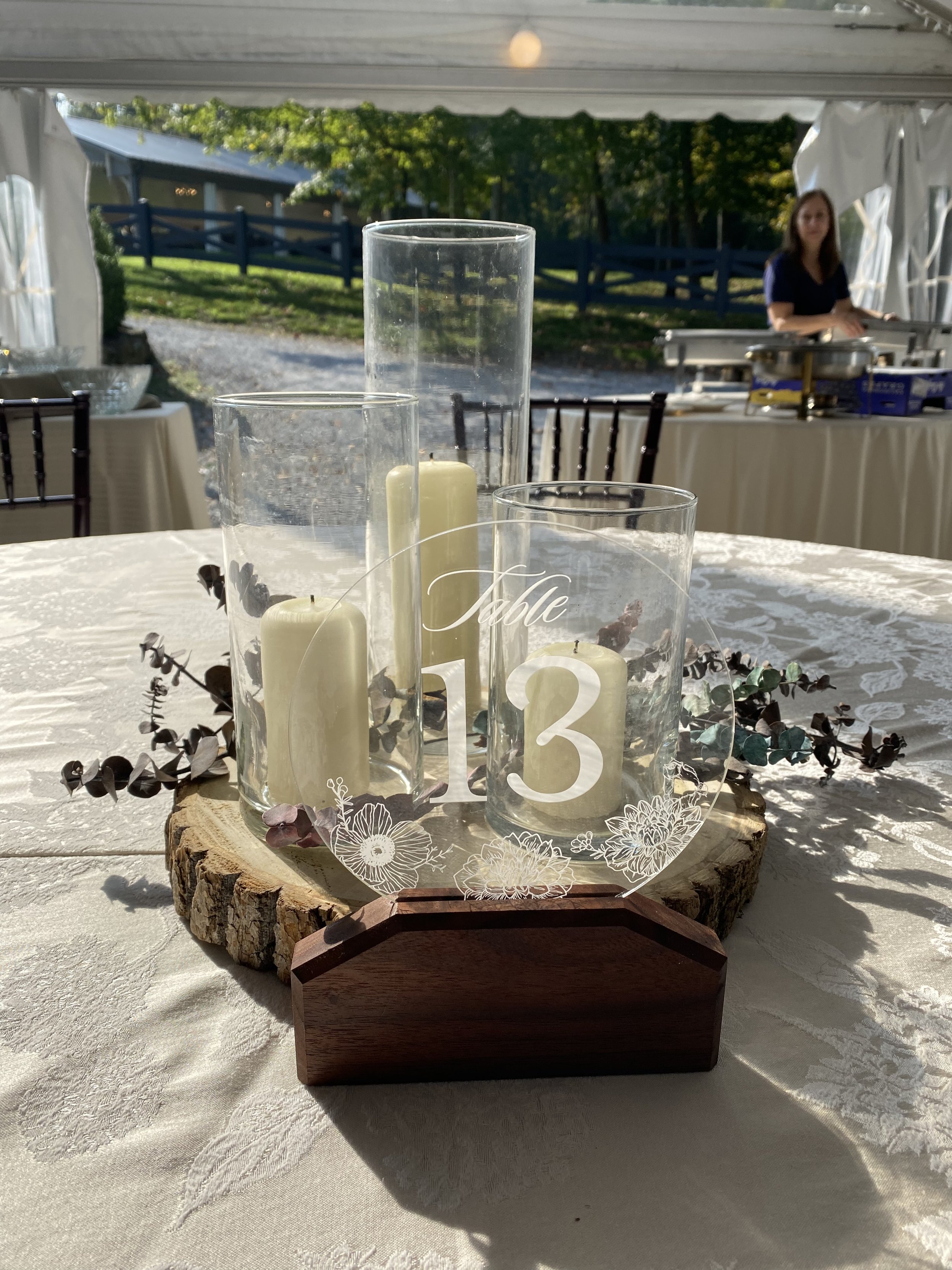 A centerpiece with white candles in glass holders and decorative greenery on a wooden slab at an outdoor event, with tables and chairs in the background.