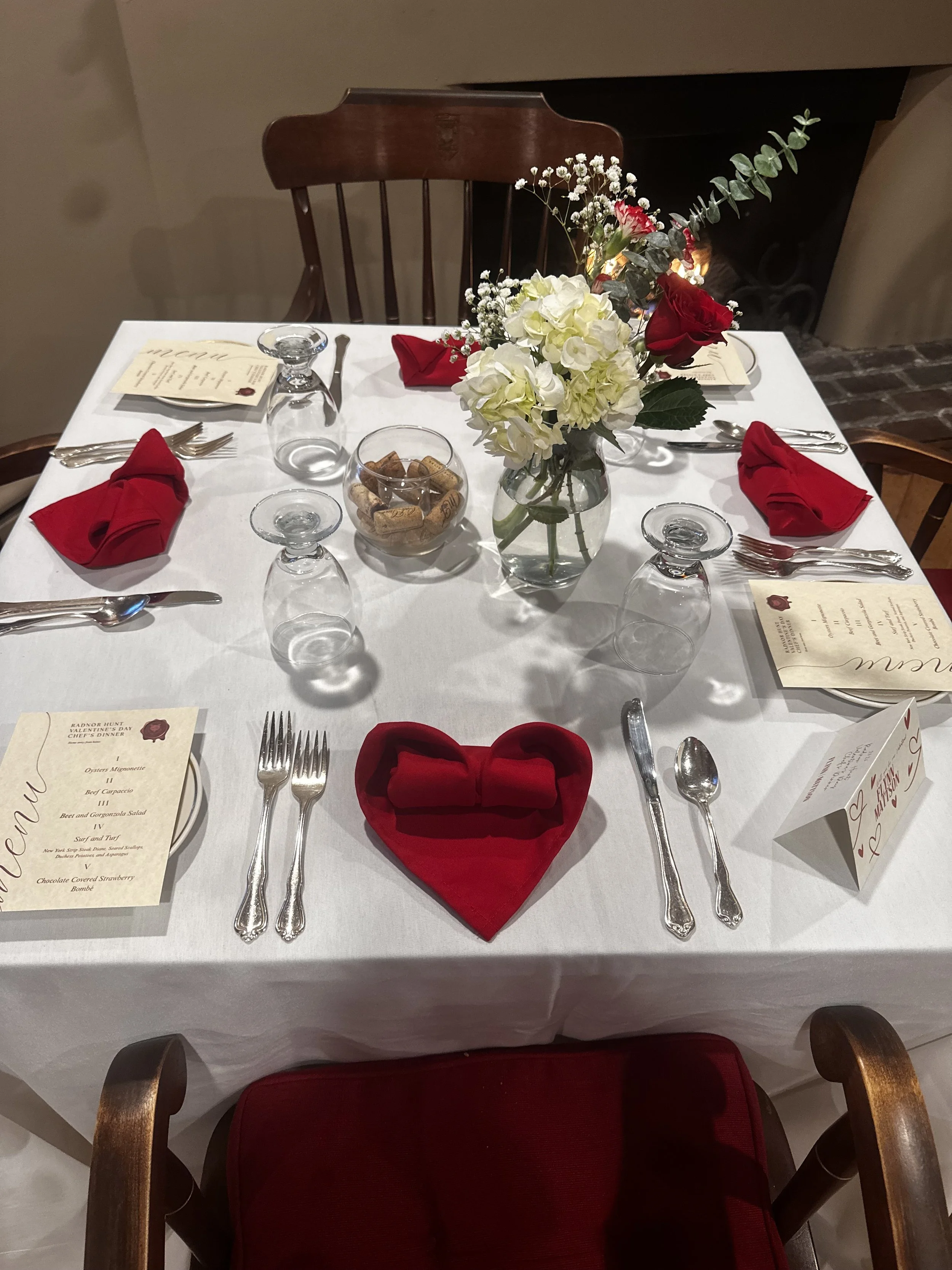 A dining table set for a meal with silverware, wine glasses, red cloth napkins, a floral centerpiece with white and red flowers in a glass vase, and menus, with dark chairs around it.