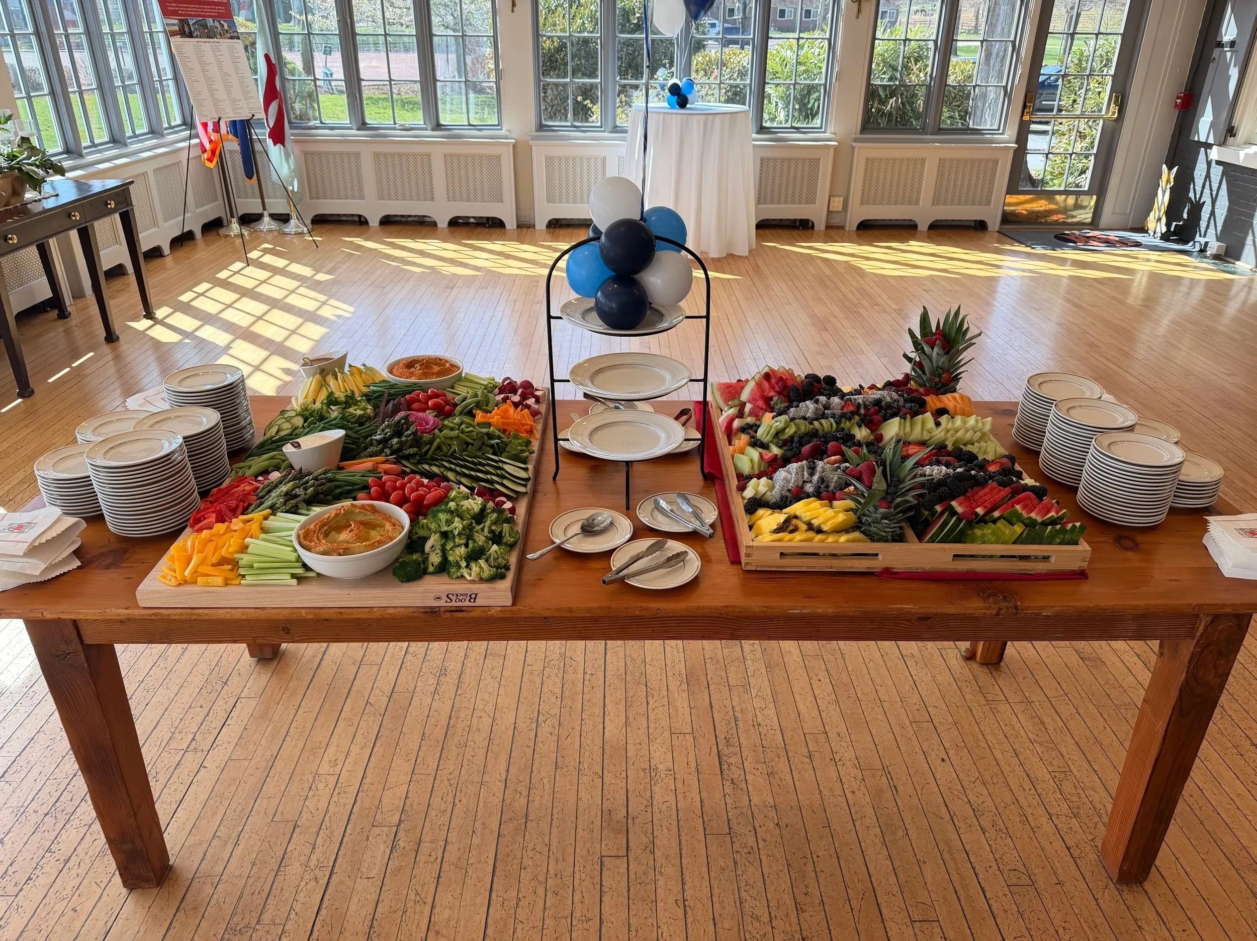 A table set up for a buffet with an assortment of fresh fruits and vegetables, plates, and utensils, in a bright room with large windows and wooden floors.