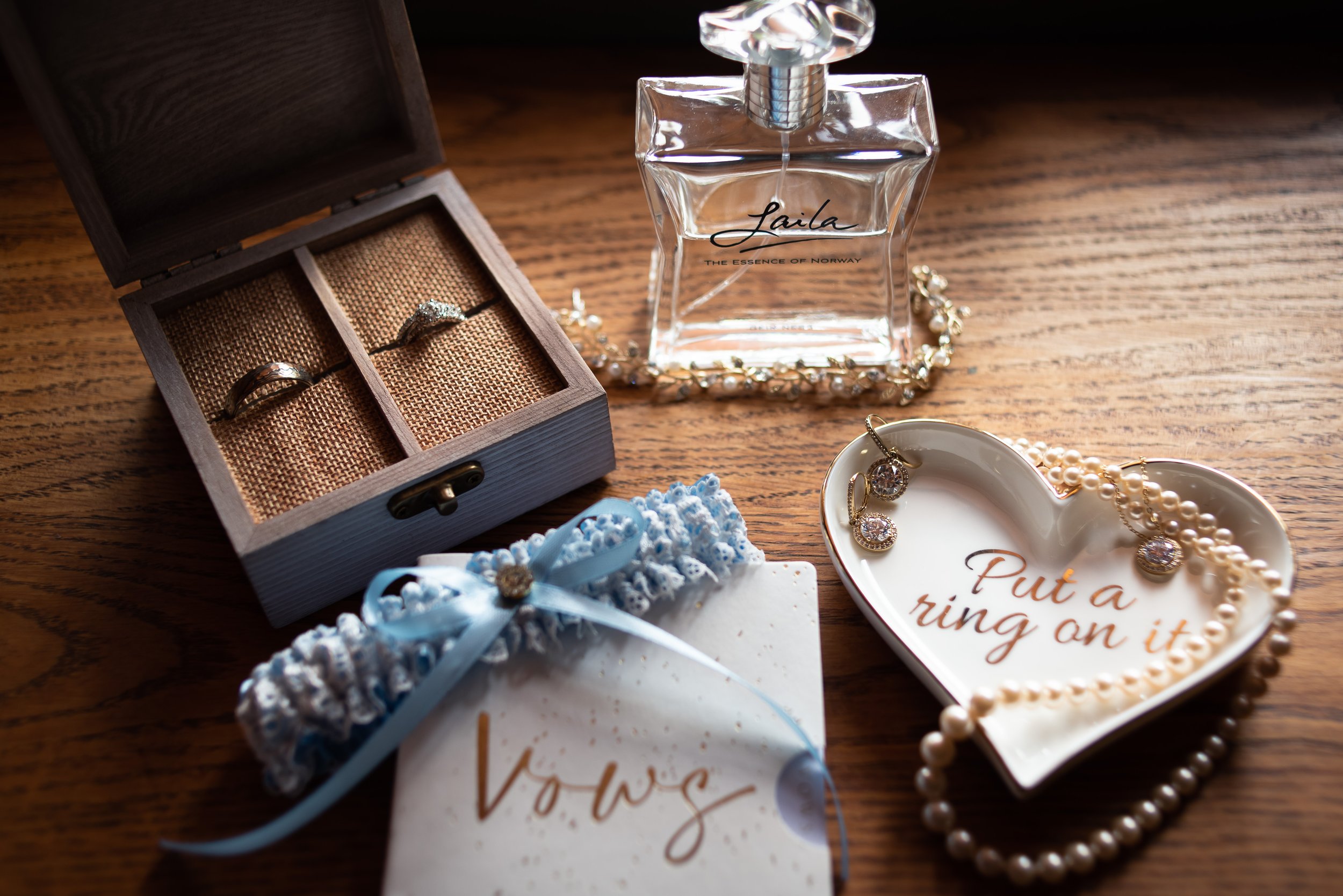 A display of jewelry and perfume on a wooden surface, including rings, earrings, pearls, a diamond ring in a wooden box, and a bottle of perfume labeled 'Laila' with the tagline 'The essence of Norway'.