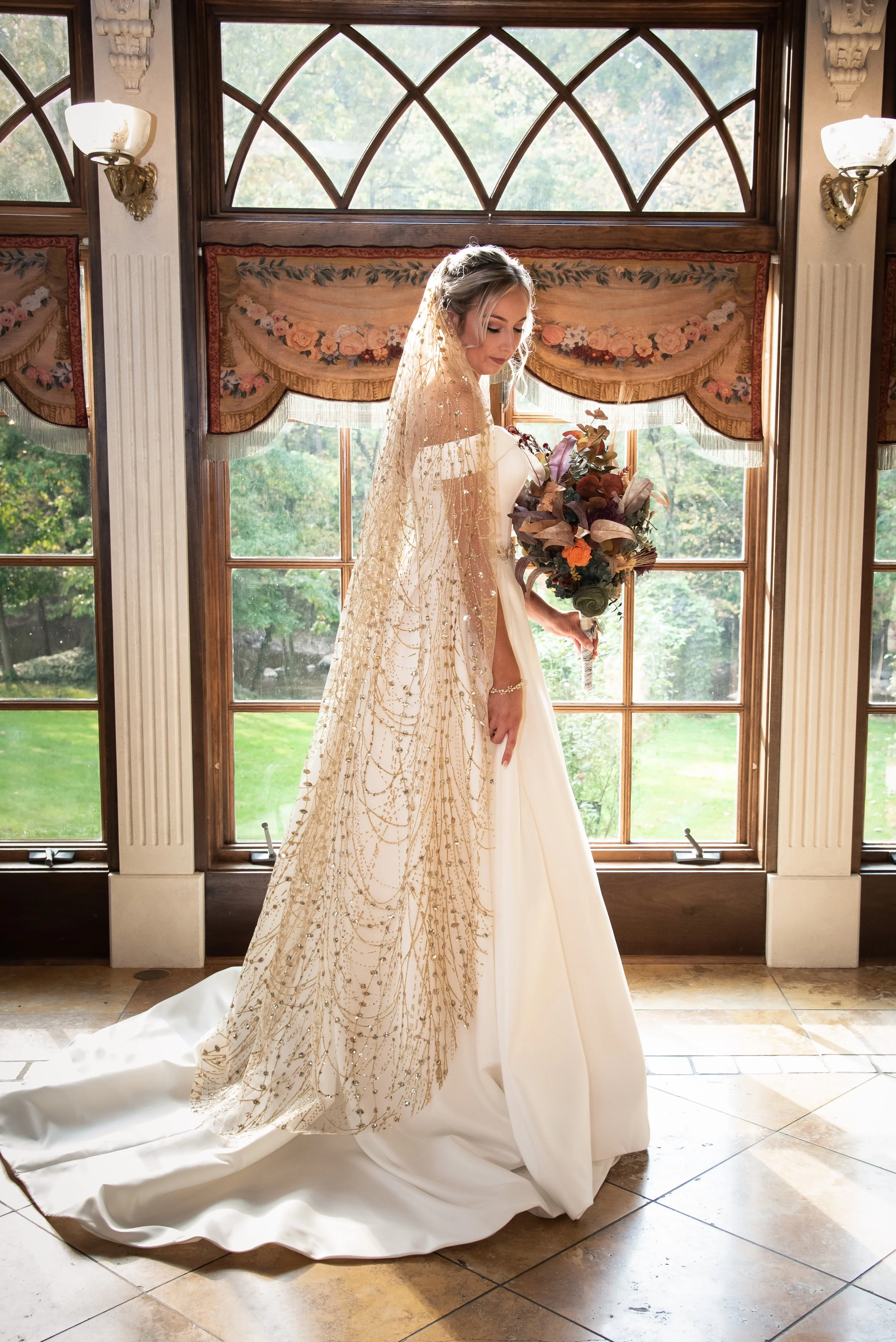A bride in a white wedding gown with gold embroidery, standing by a large window with decorative curtains. She holds a bouquet of flowers in front of her, with her veil draping over her shoulders. The sunlight streams through the window, illuminating her and the room's ornate interior.