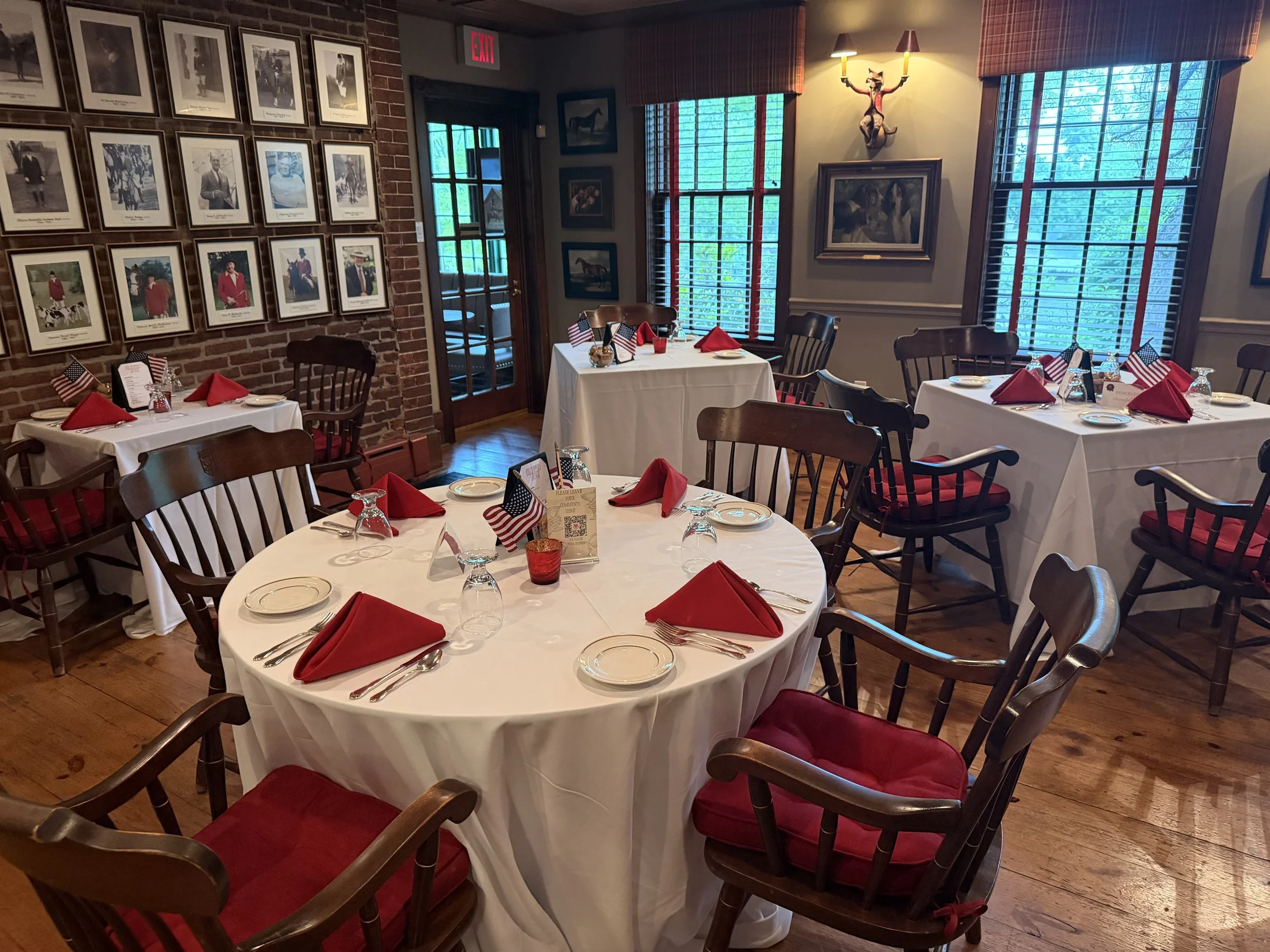 Dining room with round tables covered in white tablecloths, red napkins, American flags, and glassware, surrounded by dark chairs with red cushions. Walls decorated with framed photos and artwork, large windows with blinds, and a brick wall with a mounted animal sculpture.