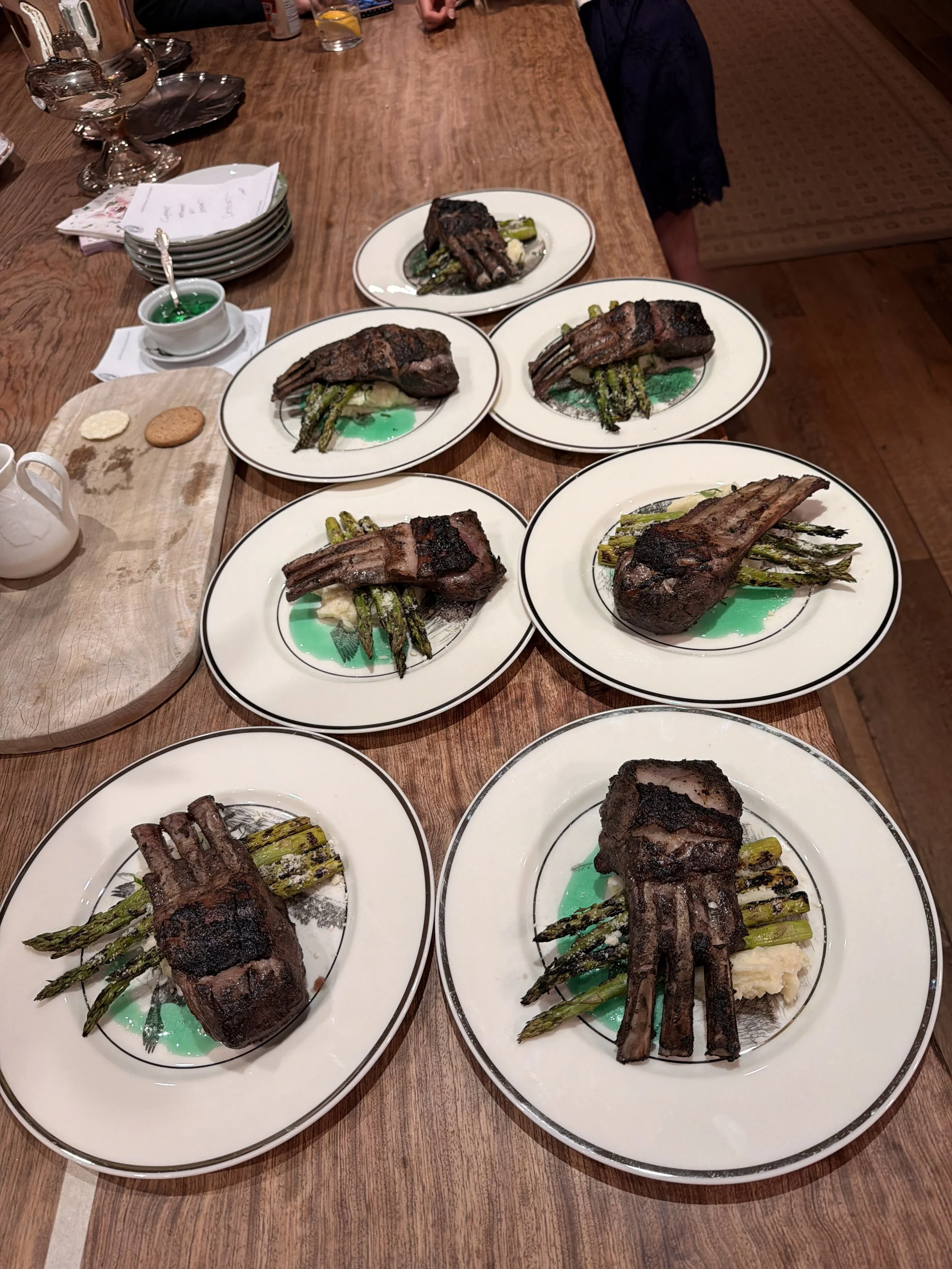 Plates with grilled lamb chops and grilled asparagus on a wooden table. There are multiple plates, some with green sauce underneath the lamb chops. In the background, there is a small plate with two cookies and a jar of peppermint candy. Also visible are glassware, napkins, and some hands and parts of people at the table.