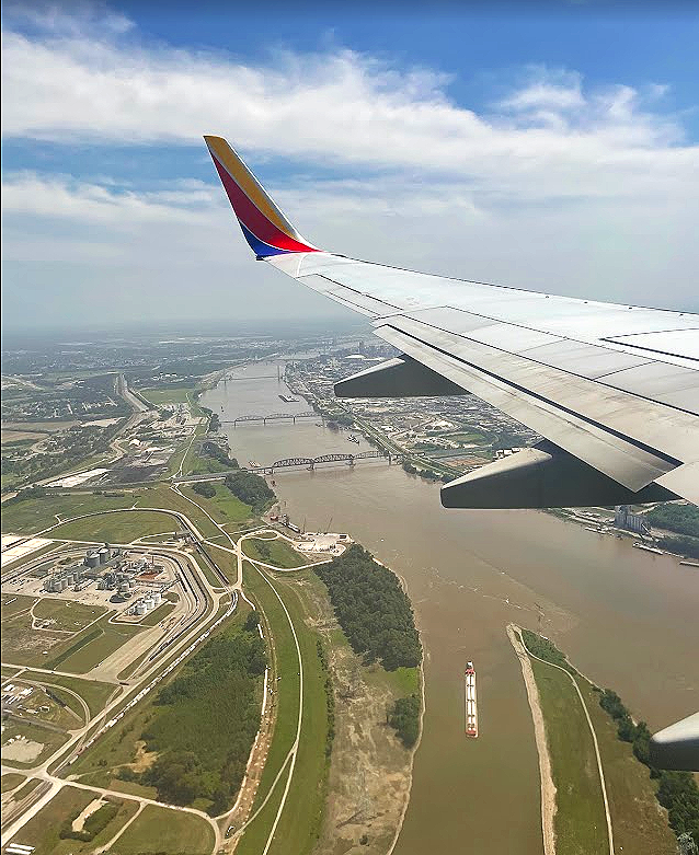 View from an airplane window showing an airplane wing over a river with bridges, green areas, and an industrial zone below.