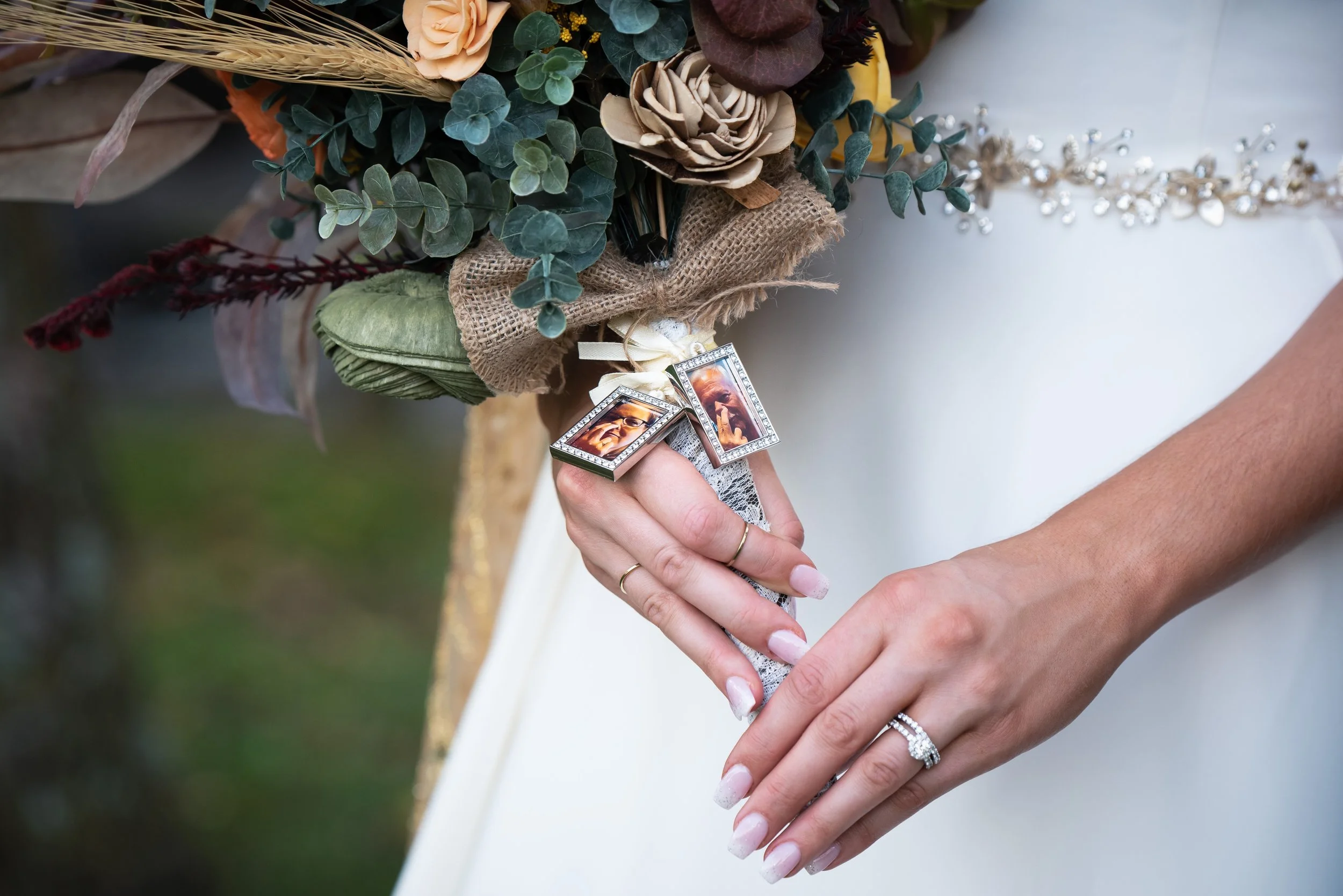A person in a wedding dress holds a bouquet of flowers wrapped in burlap. The bouquet includes greenery, peach roses, and wheat stalks. The person is wearing multiple rings, including a diamond engagement ring and wedding band, and has painted nails.