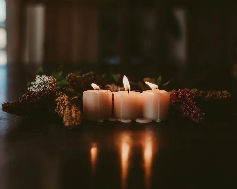 Three lit candles are arranged in a row on a dark surface, with reflections visible beneath them, surrounded by pink and white flowers.