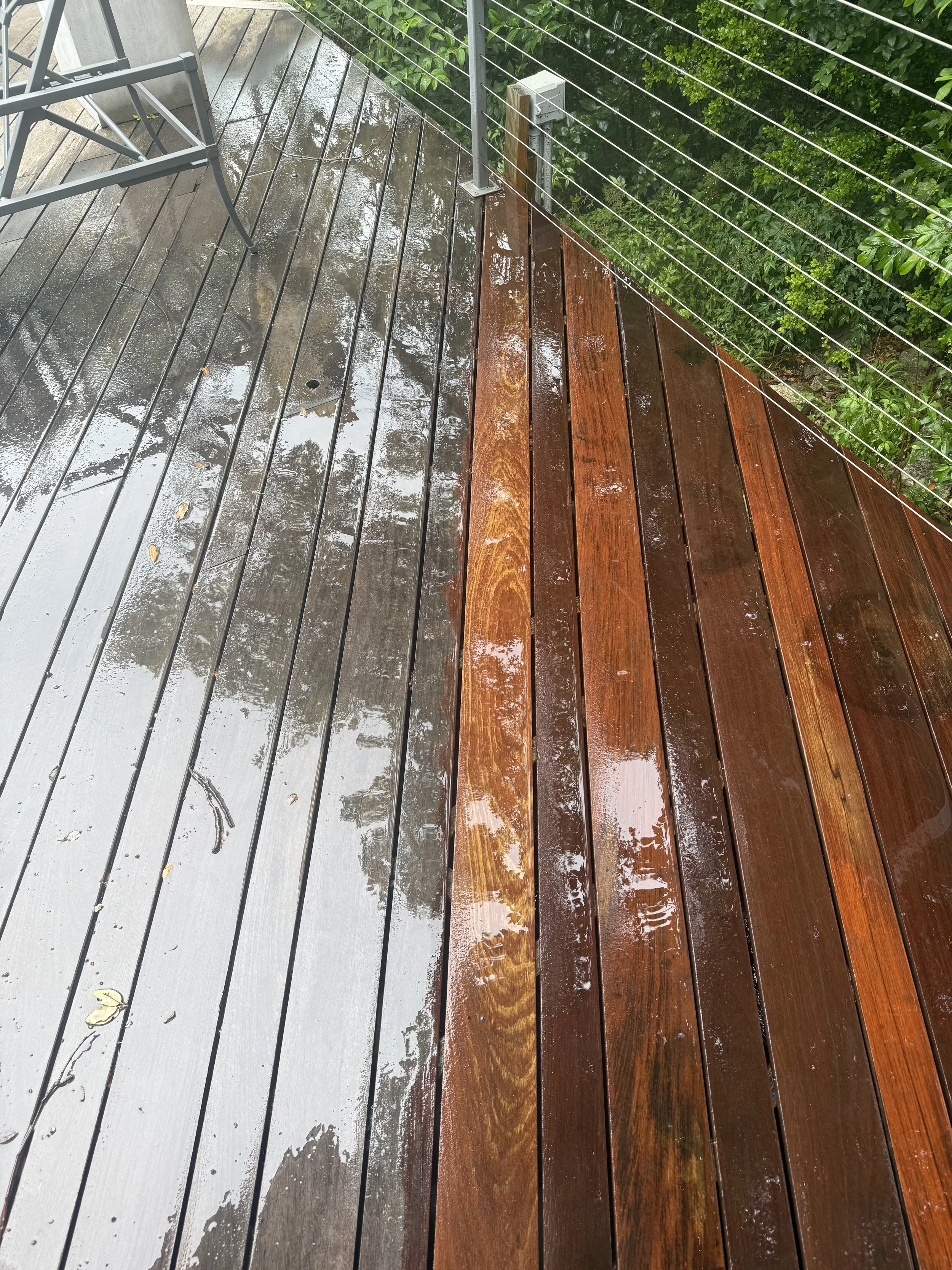 A wet wooden deck with a metal chair and a wire railing, surrounded by green bushes.