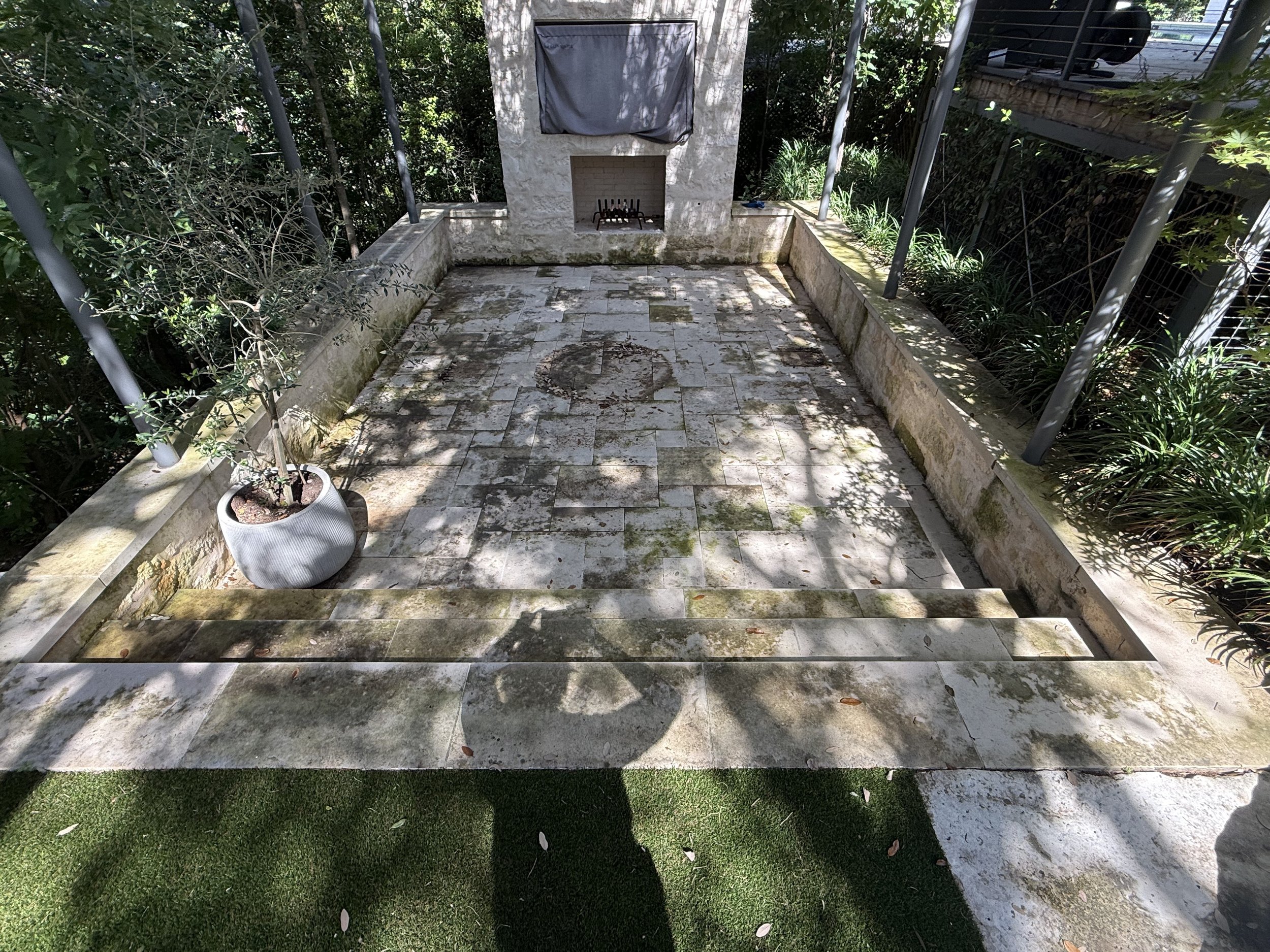 Outdoor patio with stone flooring, potted plant on left, surrounded by greenery and metal railing, shadow patterns on the ground, and a backyard with a fence and grass.