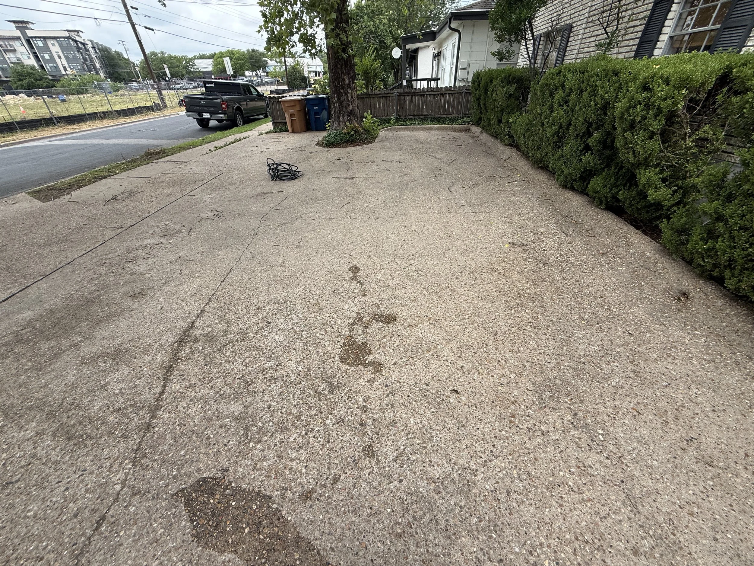 A small outdoor concrete patio with a black cord on the ground, adjacent to a small house with a brick exterior on the right, and a tall green hedge along the right side. A large tree is growing through the patio near the house, with a brown trash bin and a blue recycling bin next to it. In the background, a street with a black pickup truck, a fence, and some multi-story buildings are visible.