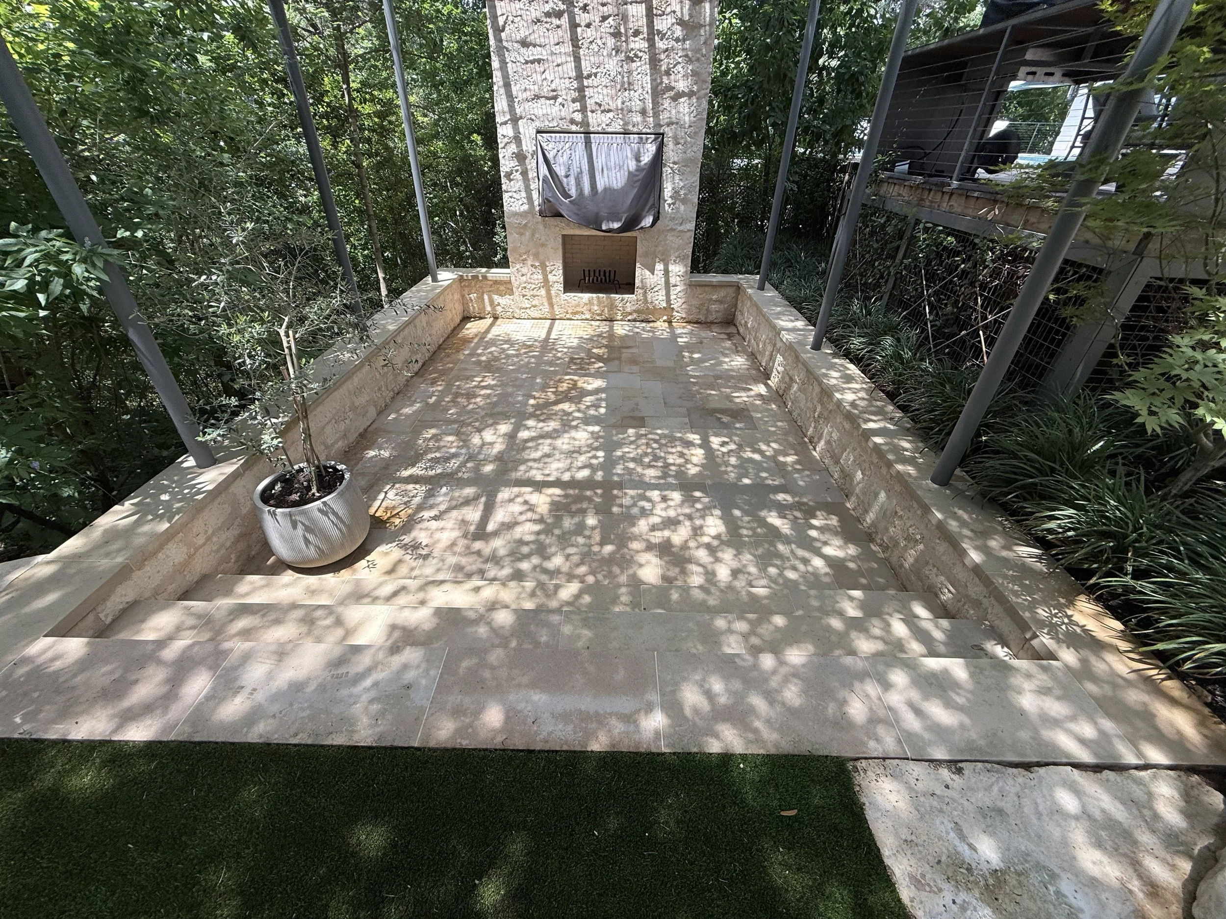 A small outdoor patio with beige stone tiles, surrounded by lush green trees and plants. There is a concrete fireplace or chimney structure in the center, covered with a gray tarp, with a black grill inside. A white planter with a small shrub is positioned at the lower left corner. Shadows of leaves cast patterns on the floor, creating a shaded and peaceful atmosphere.