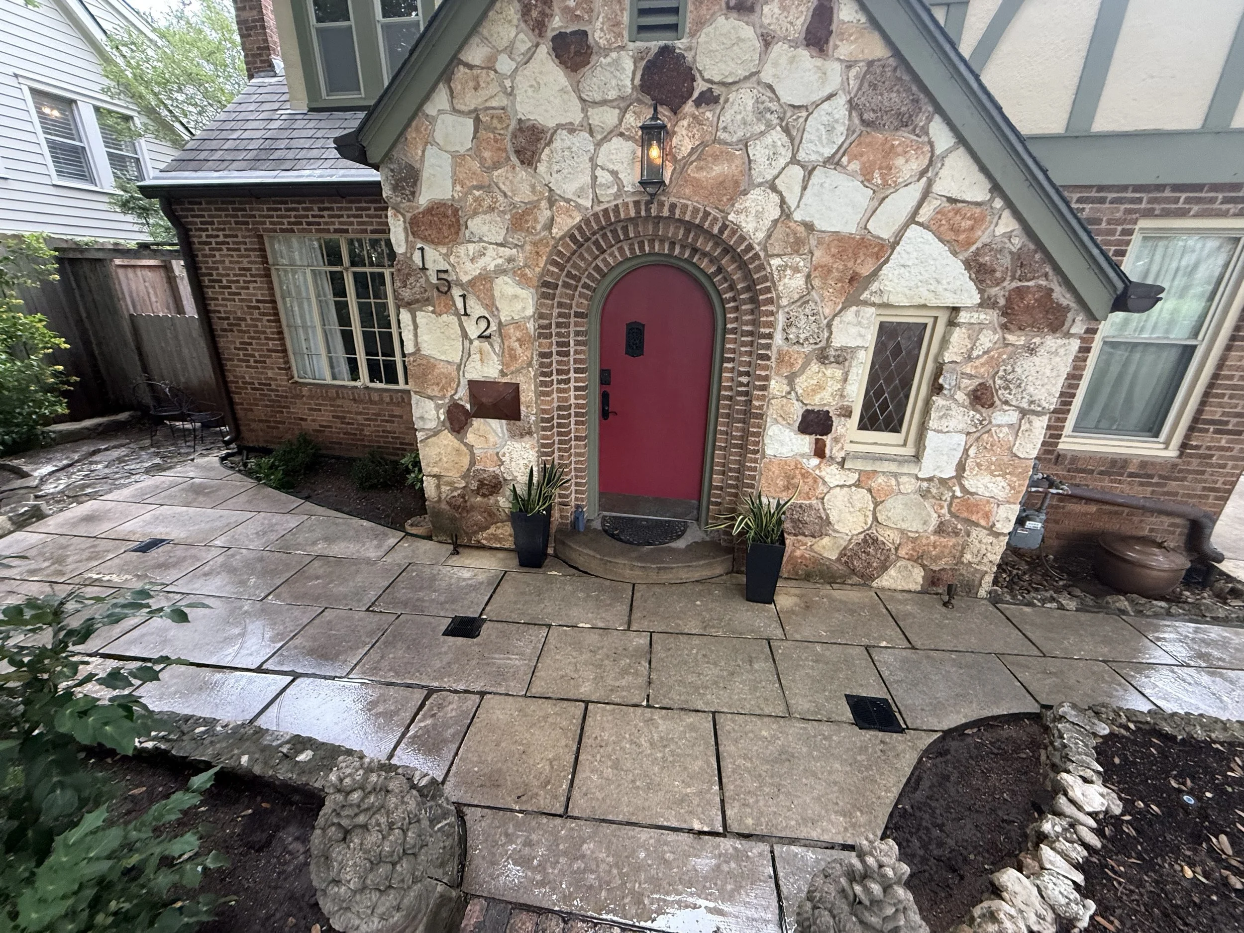 Front door with red paint, archway outlined with brick, stone house exterior, black lantern light fixture, decorative black house numbers 1512 on stone wall, potted plants in front, stone patio, outdoor seating area with chair, rain-soaked ground.