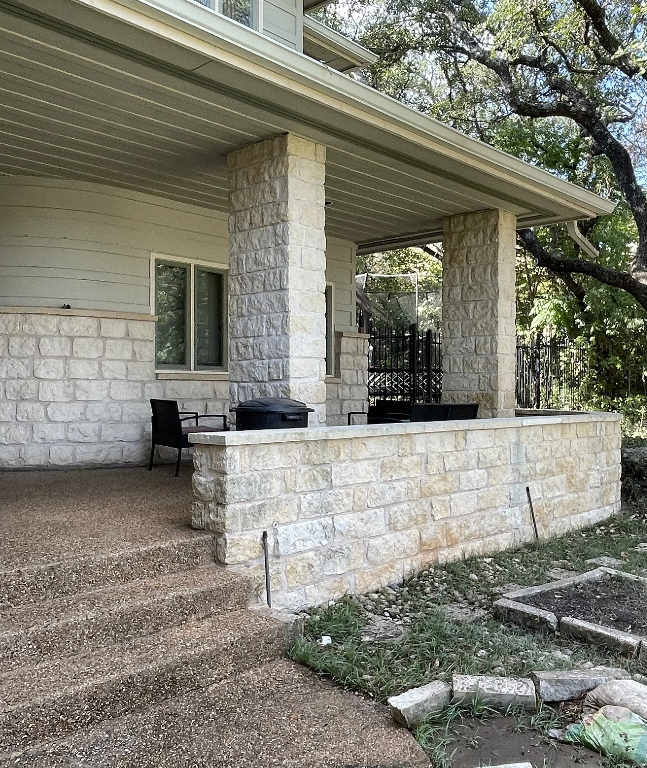 The image depicts the exterior of a house with a stone porch, supporting pillars, and sliding glass doors. There are chairs, a trash bin, and garden tools visible near the patio area, with trees and greenery in the background.