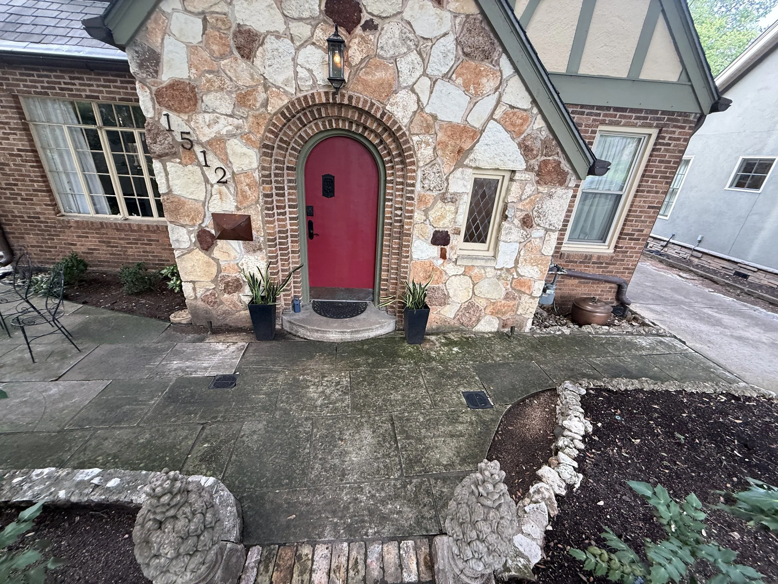 View of the front entrance of a house with a stone and brick facade. The house has a red arched door, a lantern-style light fixture above the door, and messy landscaping with rocks and soil around the garden beds. The sidewalk is wet and the scene is captured from a tilted angle.