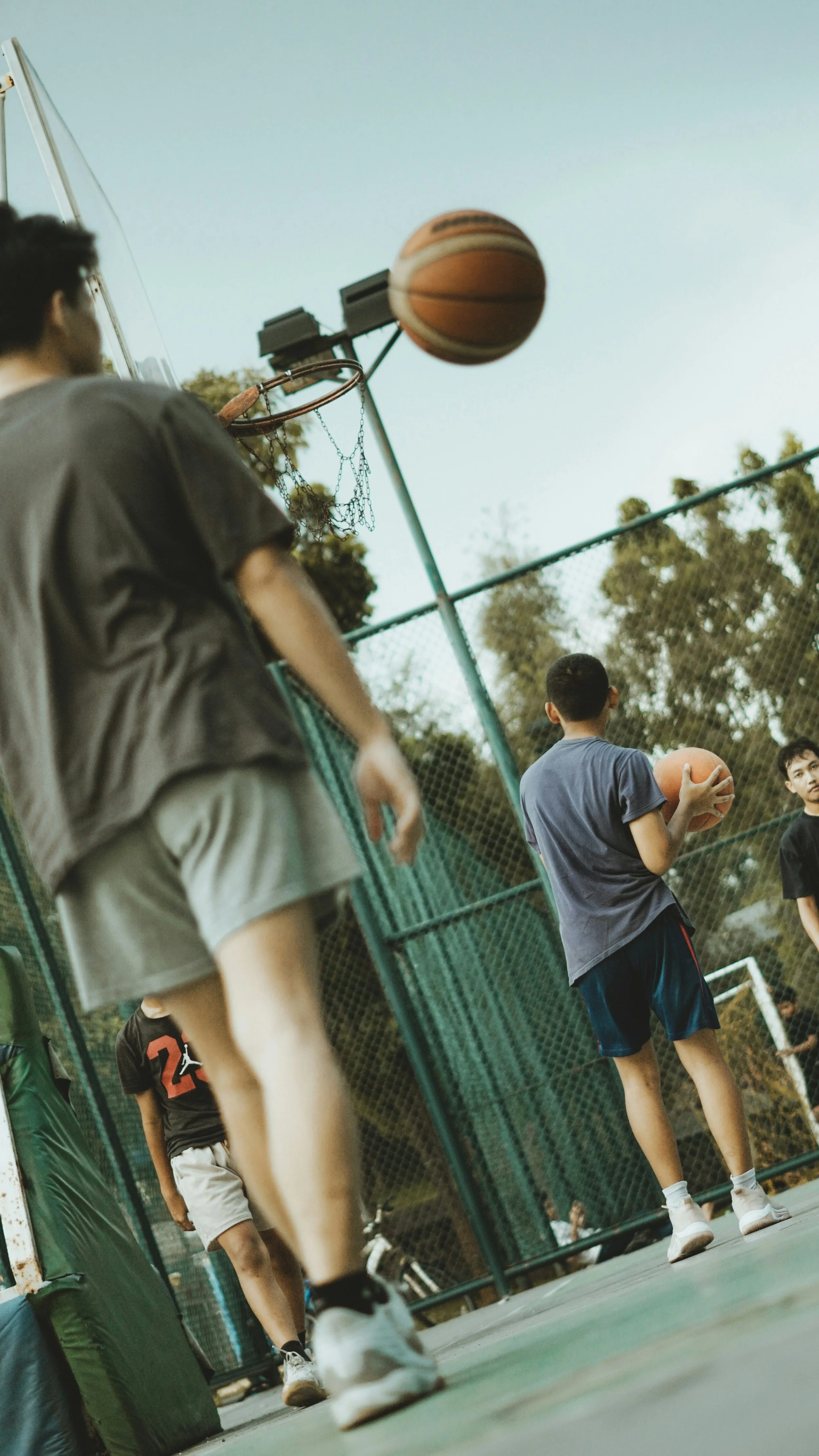 Adults playing basketball on an outdoor court with a green fence and trees in the background.