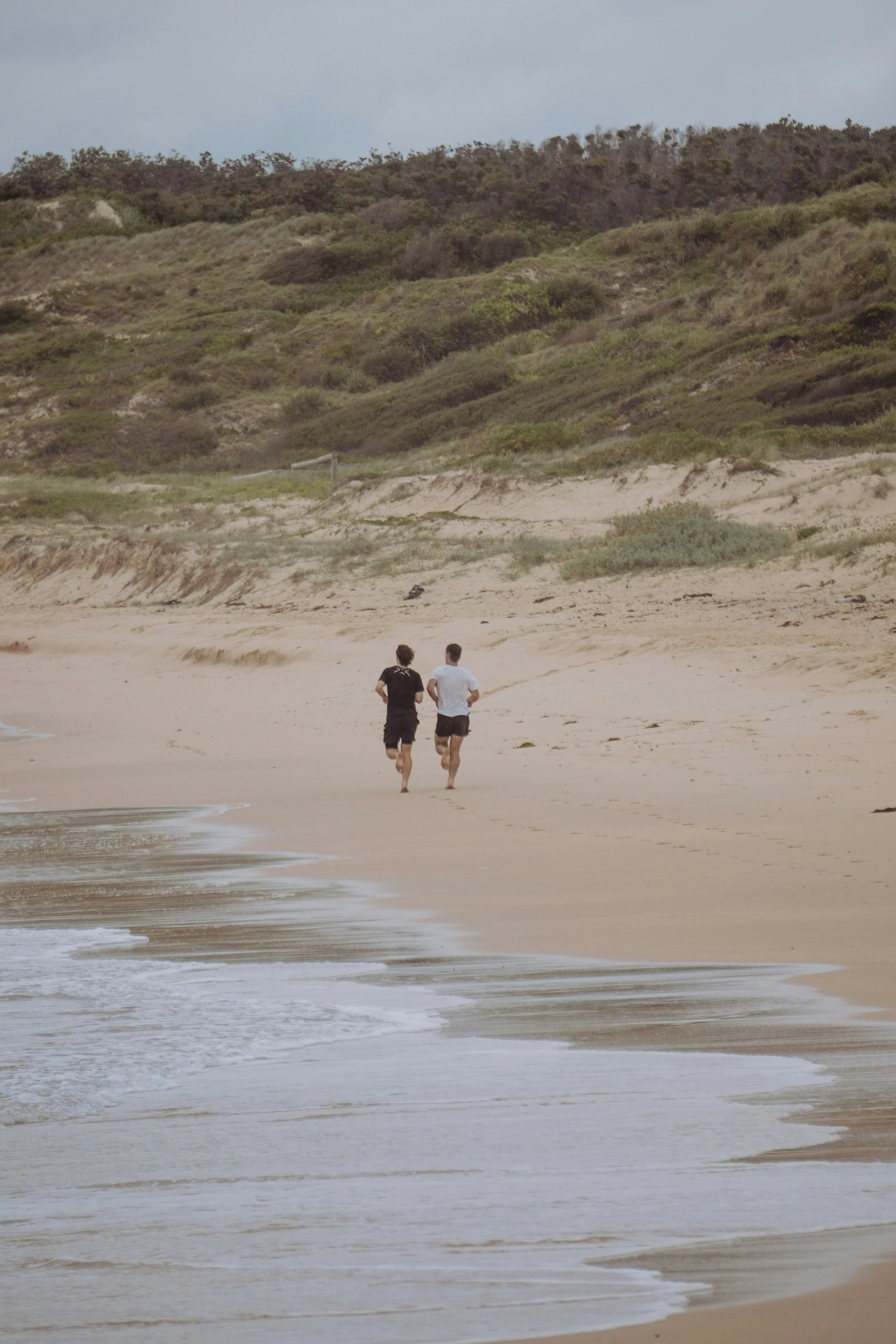 Two people jogging on a sandy beach with waves, a hill with green vegetation, and a cloudy sky in the background.