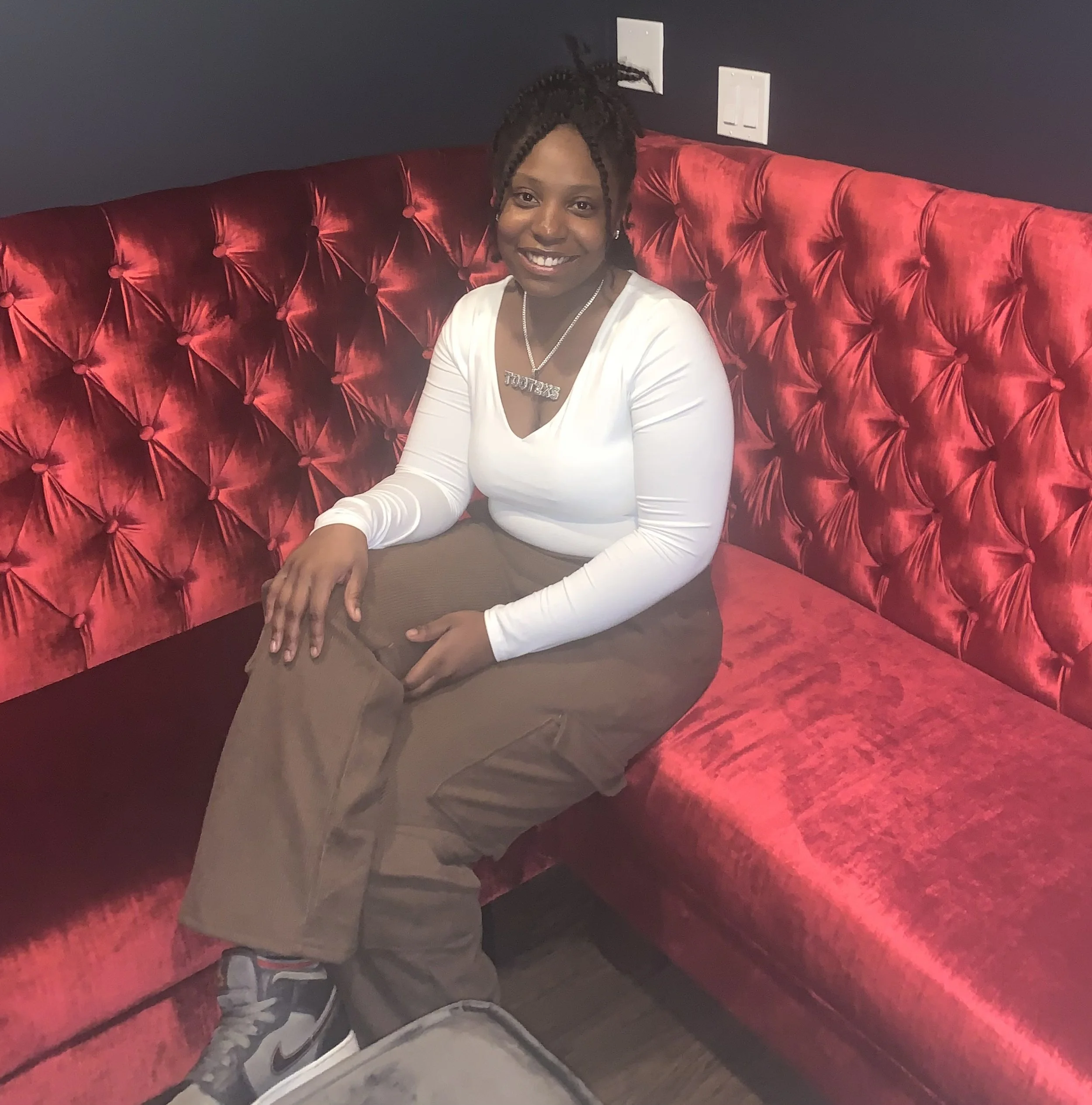 A young woman with braided hair sitting on a red velvet tufted couch, smiling at the camera, wearing a white top, beige pants, Nike sneakers, and a necklace with a pendant that says 'TooT2xs'.