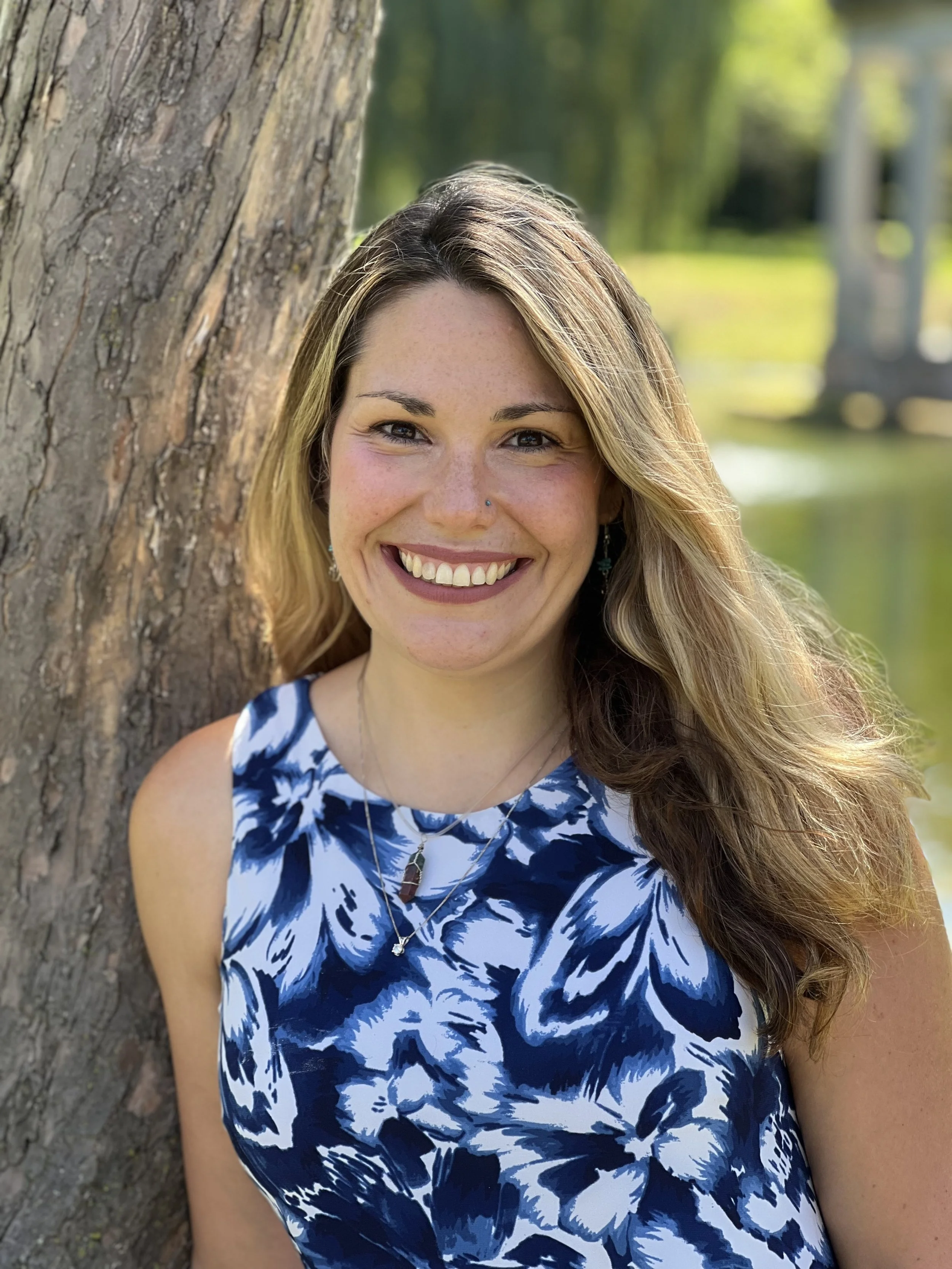 A woman with long wavy hair smiling, leaning against a tree outdoors, with a background of green grass and trees.