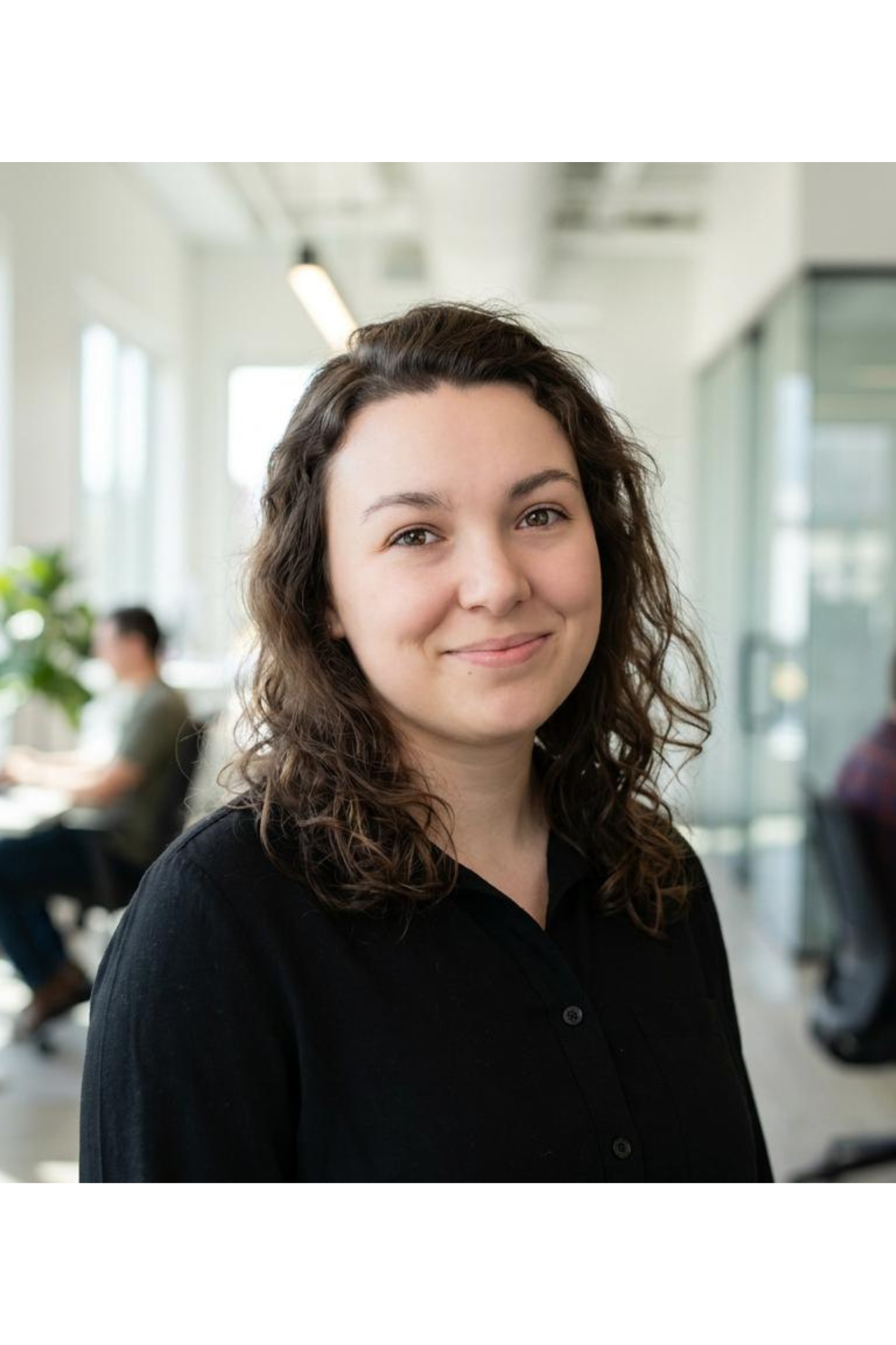 A woman with curly dark brown hair smiling in an office with large windows and people working in the background.