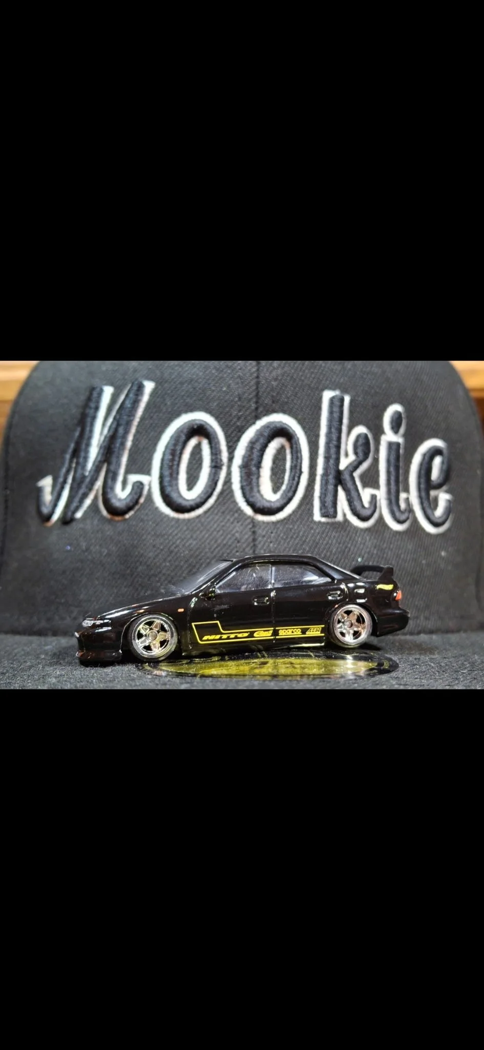 A black Toyo tires cap with embroidered lettering and a black toy car model with yellow decals, placed in front of the cap.