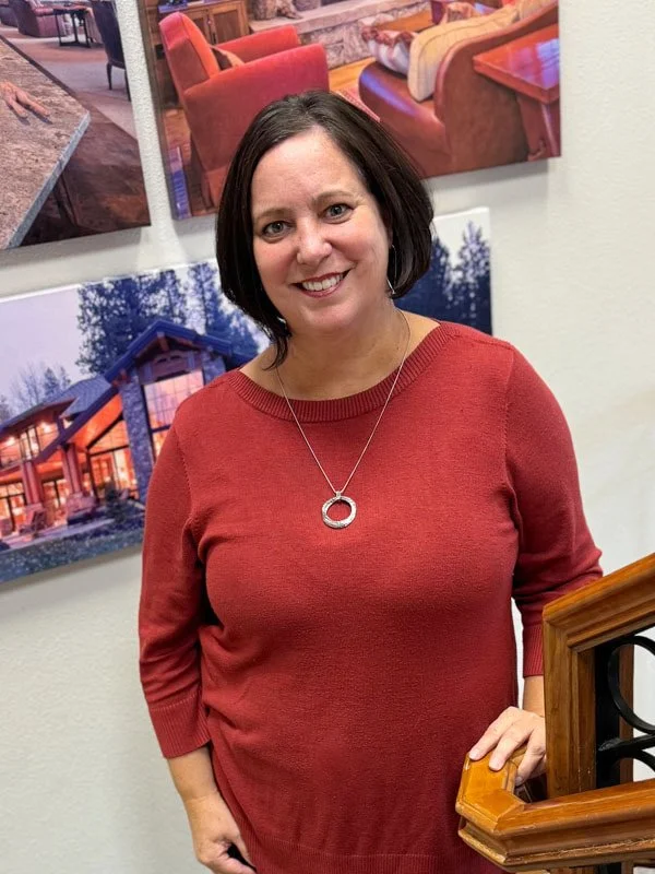 A woman with shoulder-length dark hair wearing a red sweater and silver circular pendant necklace, smiling, standing indoors in front of colorful framed artwork, holding onto a wooden railing with her right hand.