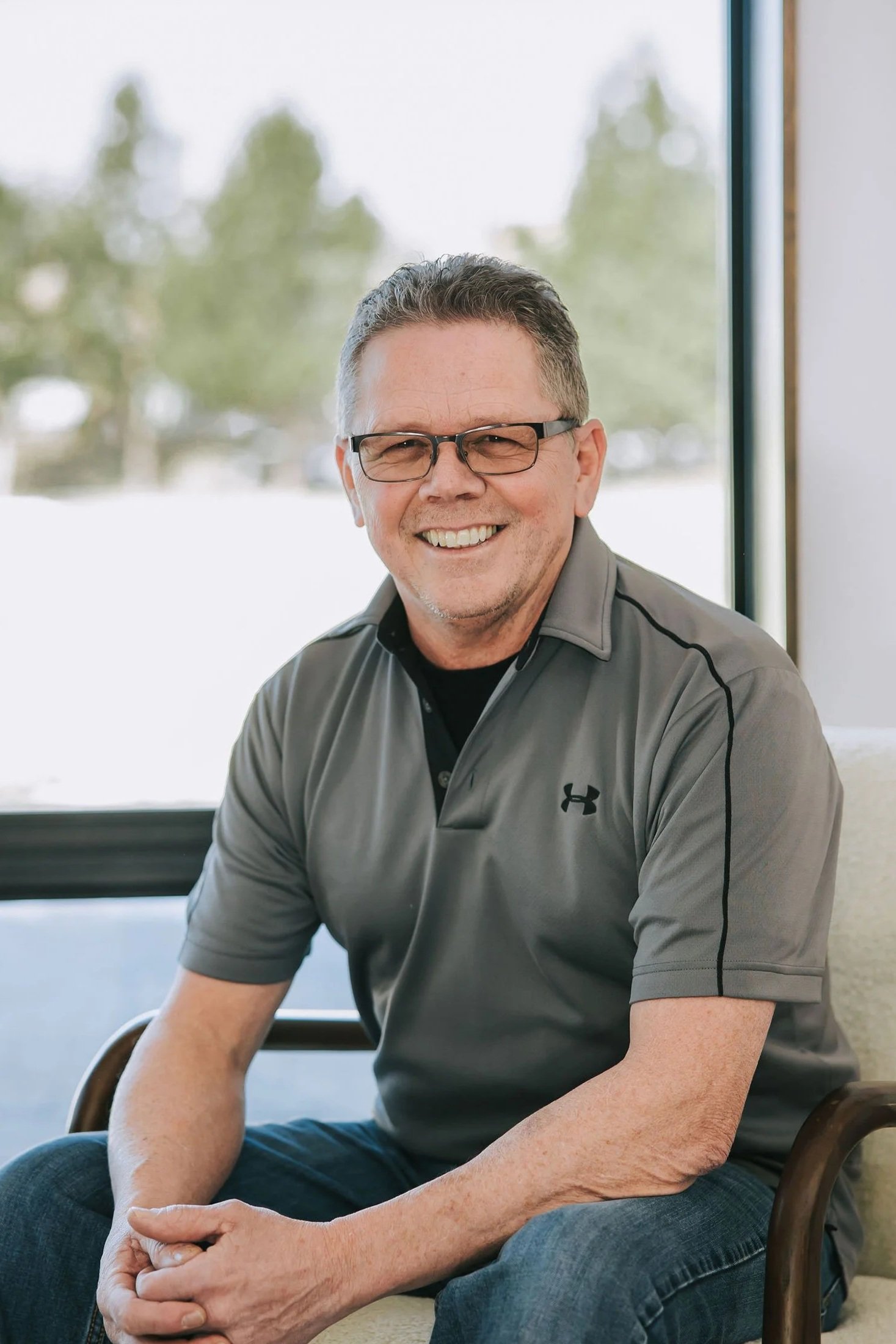 A middle-aged man with glasses smiling, wearing a gray Under Armour polo shirt, sitting indoors by a large window with blurred greenery outside.
