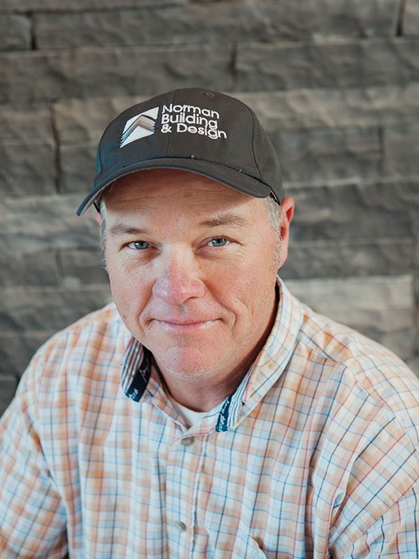 A man wearing a black hat with the logo and text 'Norman Building & Design' smiling, wearing a light plaid shirt, with a wood-textured wall in the background.