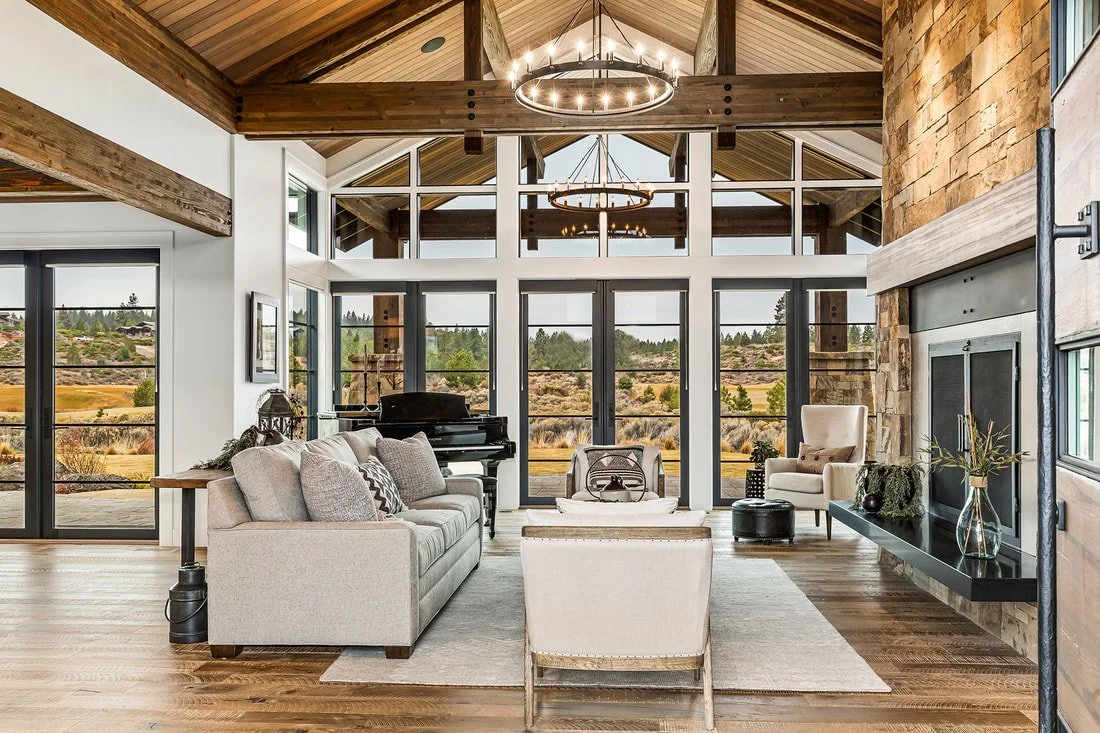 Modern living room with large windows, wooden ceiling, stone fireplace, beige sofa, armchair, and grand piano under a circular chandelier.